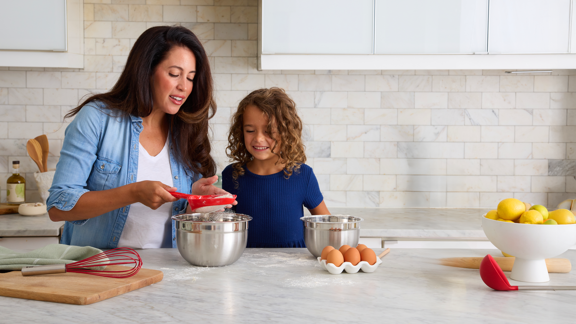 A woman and a young girl are baking together in a bright kitchen, mixing ingredients in metal bowls on a counter with eggs, lemons, and kitchen utensils nearby.