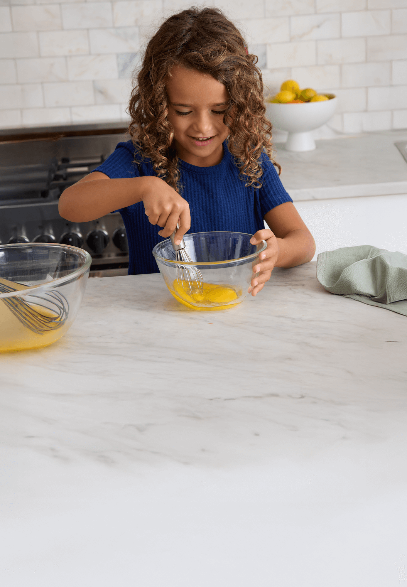 A young child with curly hair and a blue shirt is whisking eggs in a glass bowl on a kitchen counter. Another bowl with a whisk, a towel, and a bowl of lemons are on the white countertop in the background.