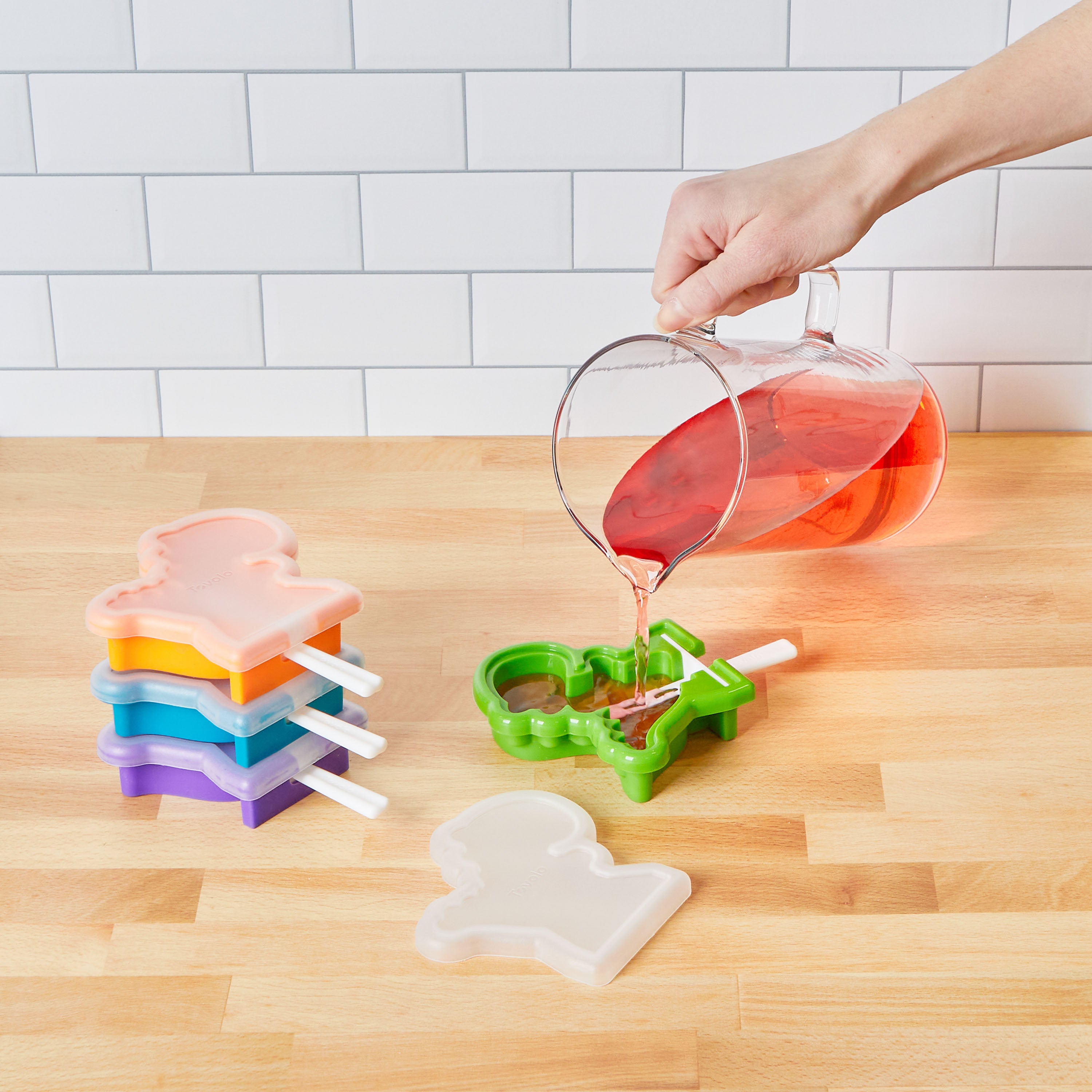A hand pours red liquid from a clear pitcher into a green Pop Molds Stackable popsicle maker on a wooden surface, with four stackable pop makers and a white tiled wall in the background.