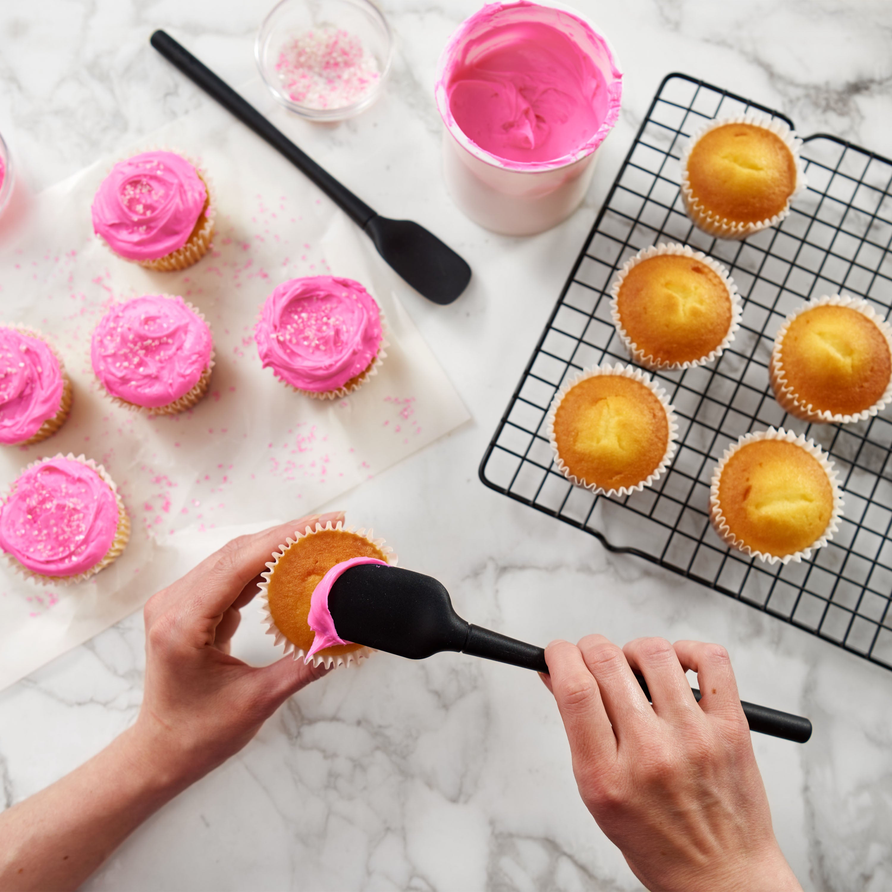 A person uses the Flex-Core Mini Spat + Spoonula to spread pink frosting on a cupcake. More frosted cupcakes with sprinkles, plain cupcakes on a cooling rack, and a jar of pink frosting sit nearby on a marble surface.
