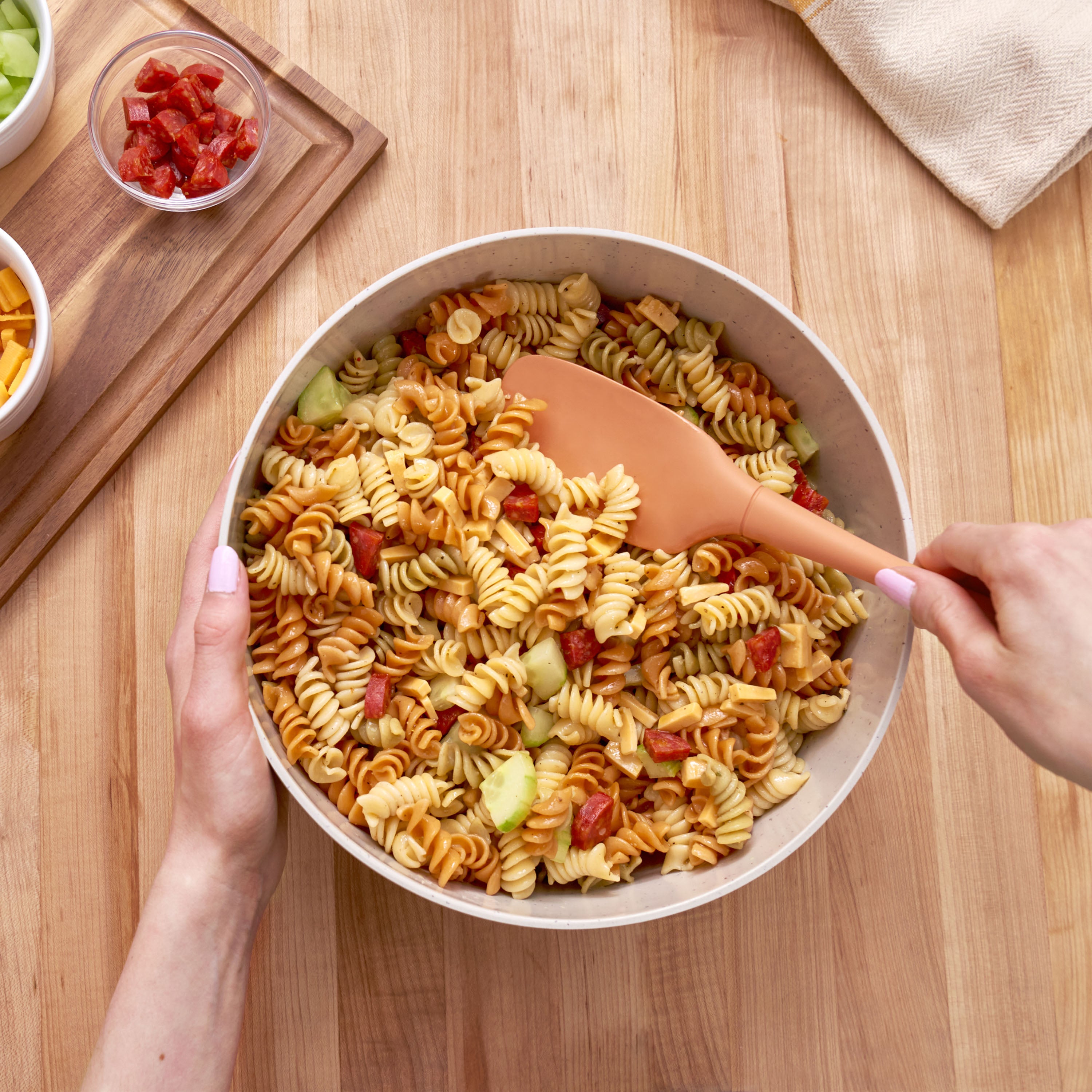 A person mixes pasta salad with rotini, diced vegetables, and pepperoni in a large bowl using a Flex-Core Spoonula on a wooden table, with small bowls of extra ingredients nearby.