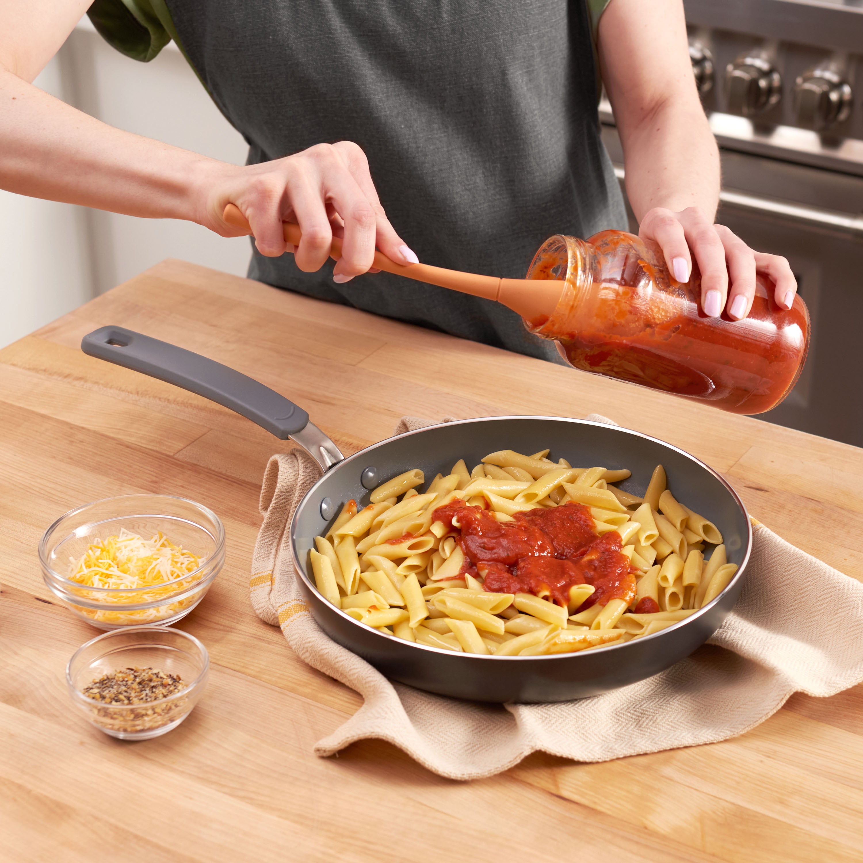 A person uses the Flex-Core Jar Scraper to pour tomato sauce from a jar onto cooked penne in a skillet on a wooden counter, with bowls of shredded cheese and herbs nearby.