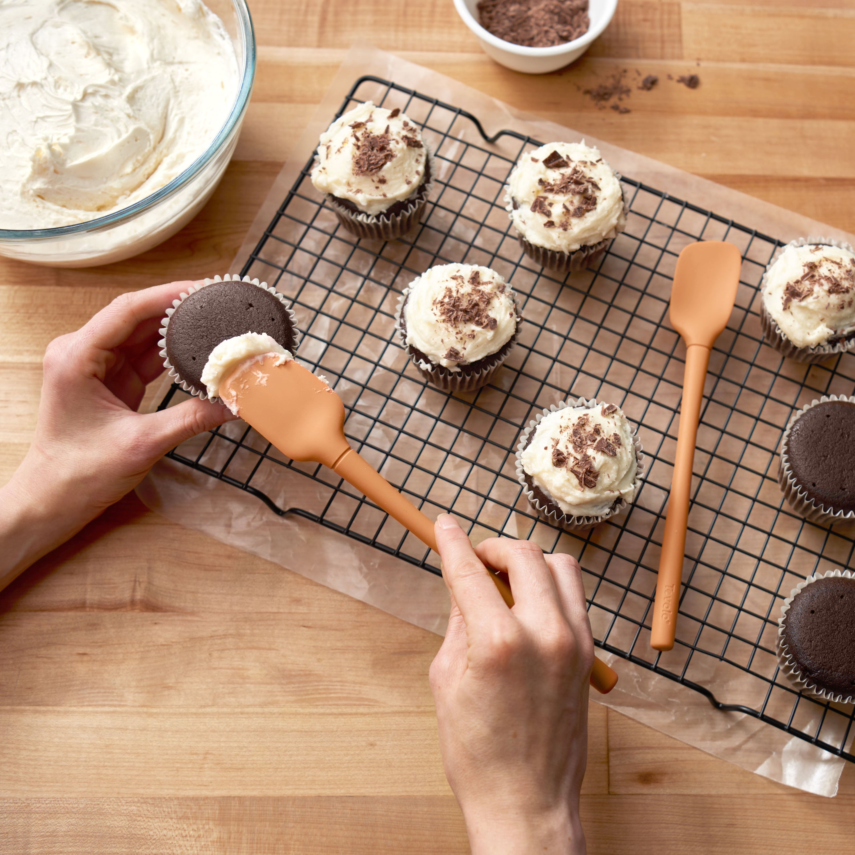 Using the Flex-Core Mini Spat + Spoonula, a person frosts chocolate cupcakes with white icing on a cooling rack. Nearby are more cupcakes, a bowl of frosting, and chocolate shavings on a wooden table.