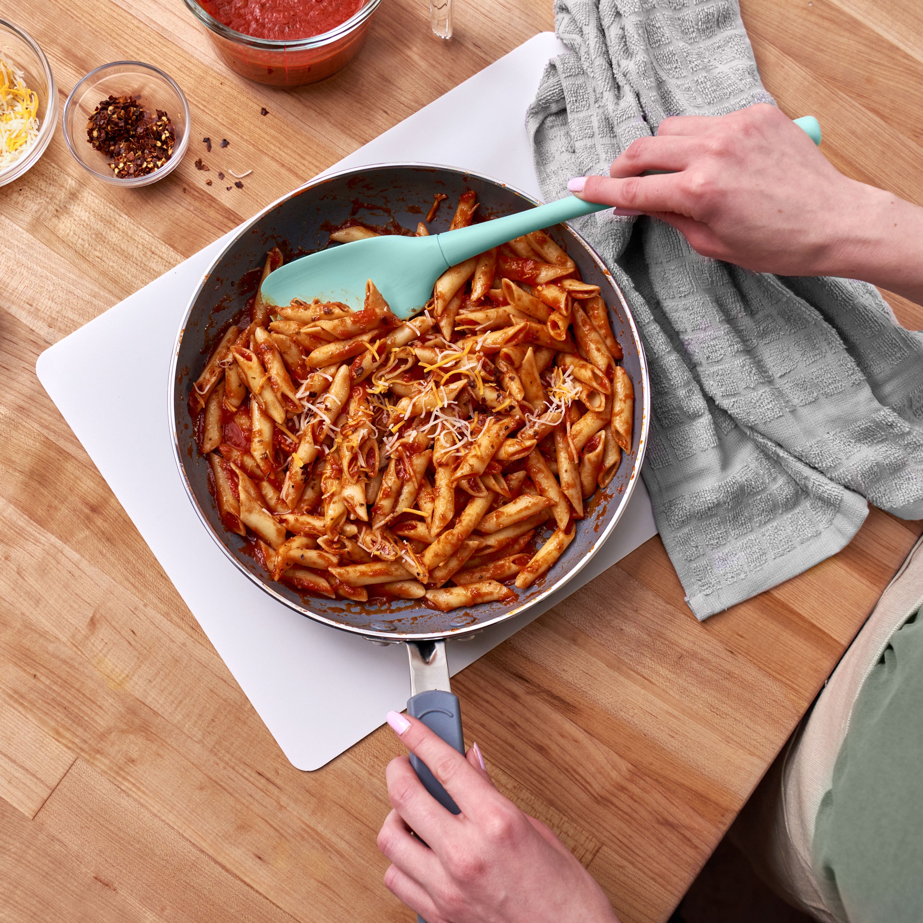 A person stirs penne pasta with tomato sauce and shredded cheese in a frying pan, using the teal Flex-Core Spoonula. On the wooden countertop are a gray dish towel and small bowls of sauce, cheese, and red pepper flakes.