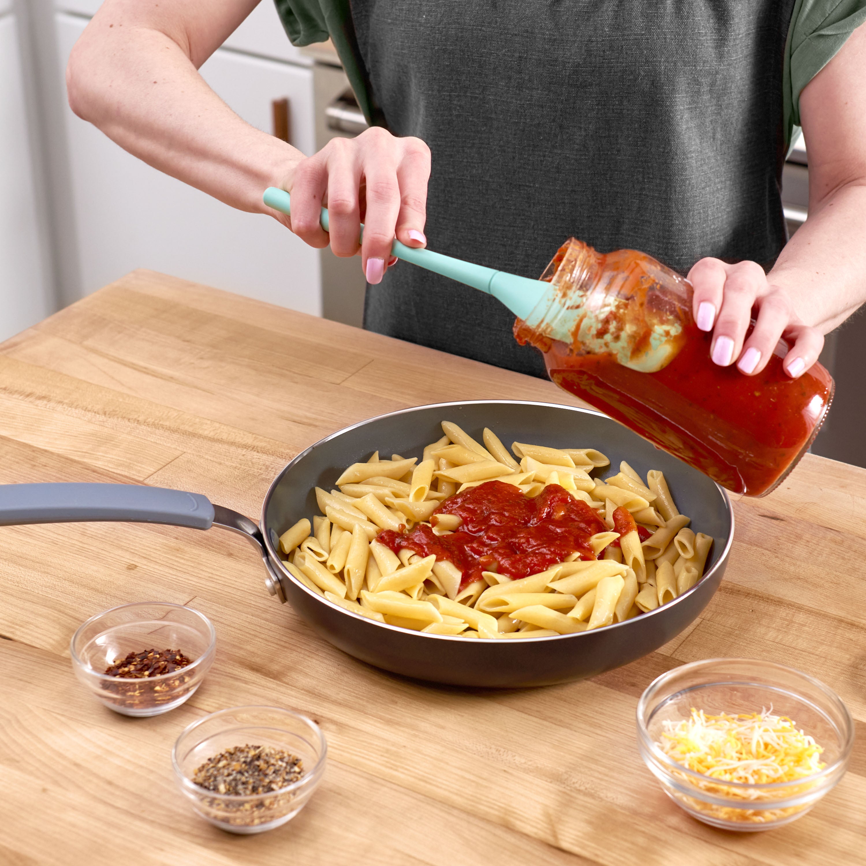 Using the Flex-Core Jar Scraper, someone pours tomato sauce from a jar onto penne pasta cooking in a skillet on a wooden countertop. Nearby sit bowls of shredded cheese, red pepper flakes, and dried herbs.