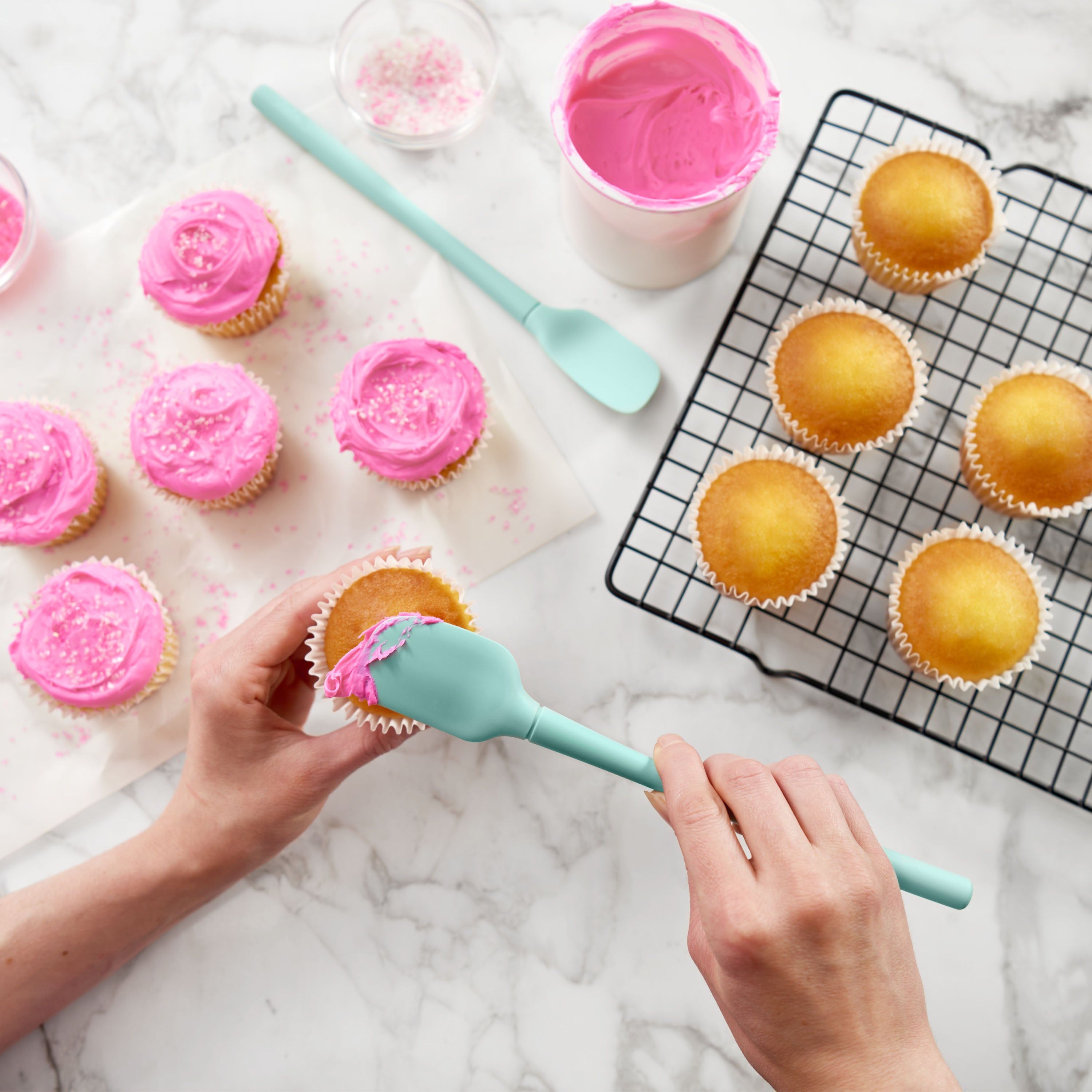 Using a Flex-Core Mini Spat + Spoonula, a person frosts yellow cupcakes with pink icing on a marble counter. Some cupcakes are on a cooling rack; others are already frosted and sprinkled. A bowl of pink frosting is nearby.