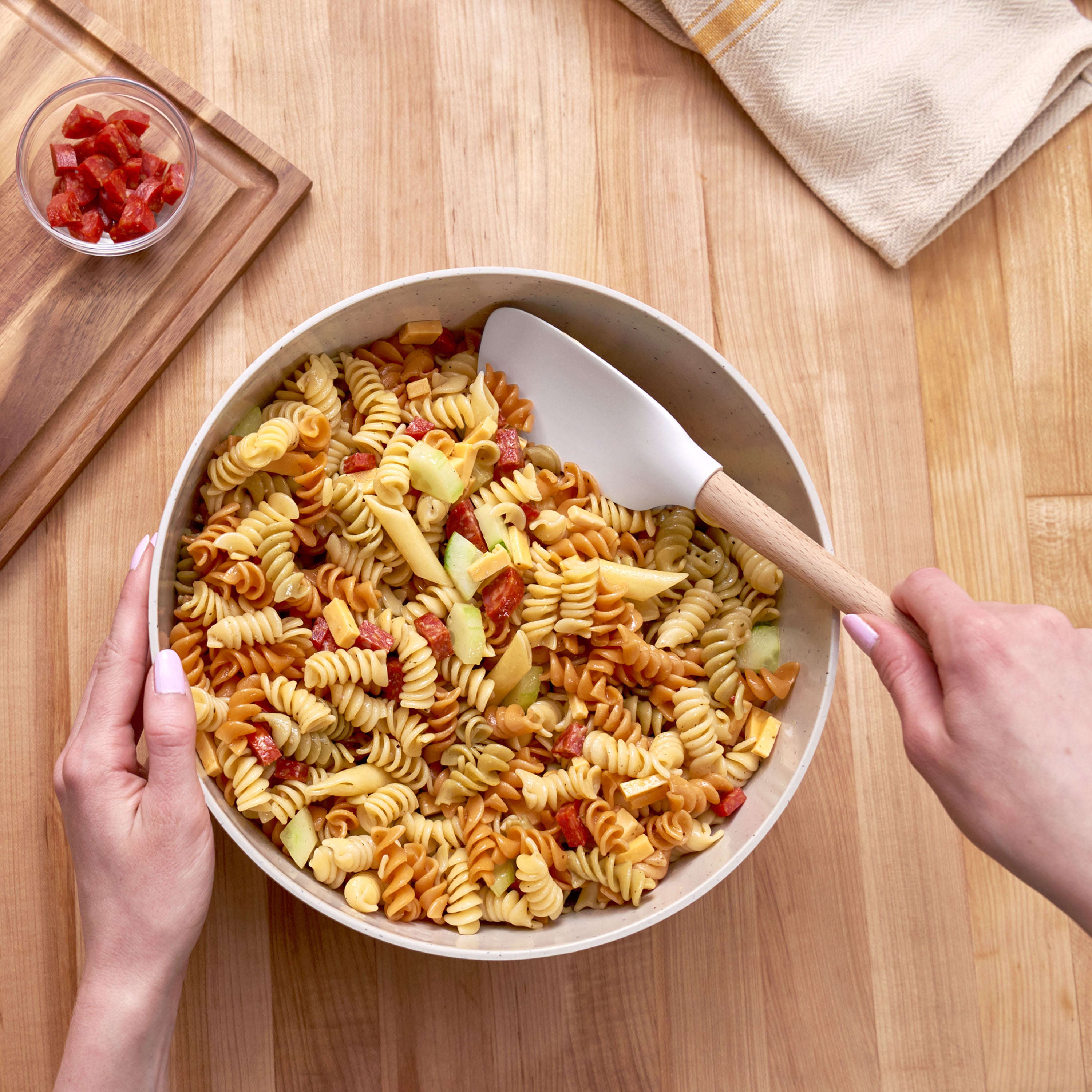 Someone mixes rotini pasta salad with sliced veggies and diced pepperoni using a Flex-Core Spoonula on a wooden table, with a small bowl of extra pepperoni nearby.