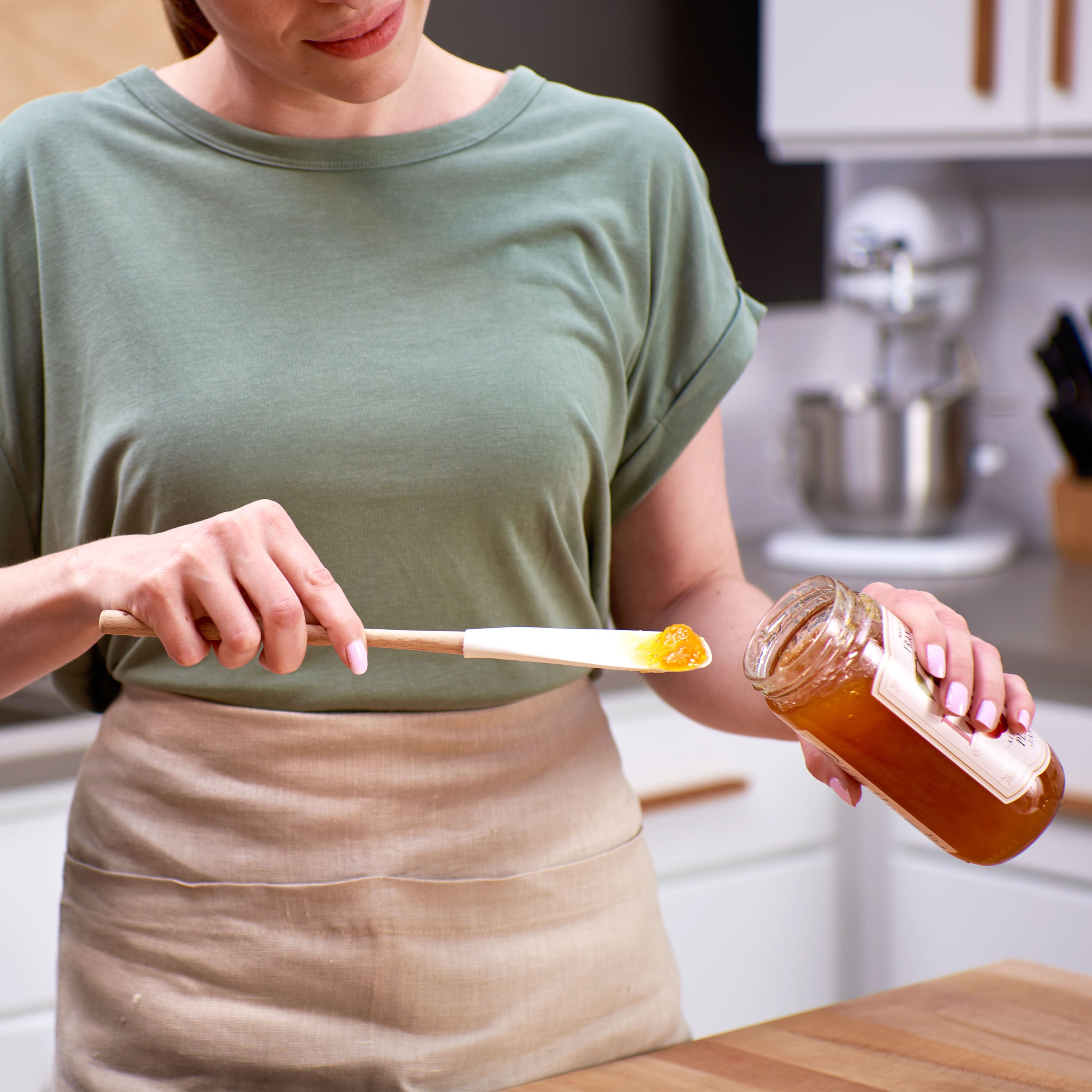 In a bright kitchen, a woman in a green shirt and beige apron uses the Flex-Core Jar Scraper to spread honey from a glass jar onto a silicone spatula.