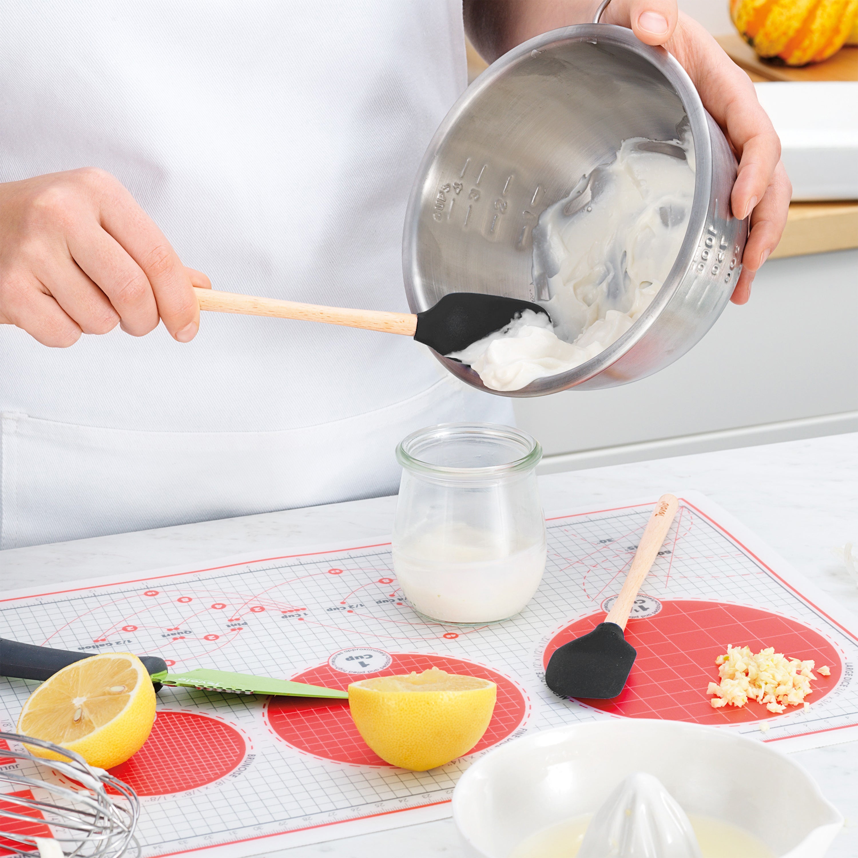 Someone uses the Flex-Core Mini Spat + Spoonula in black silicone to scrape a creamy mixture from a metal bowl into a glass jar on a kitchen counter with lemons, garlic, and utensils nearby.