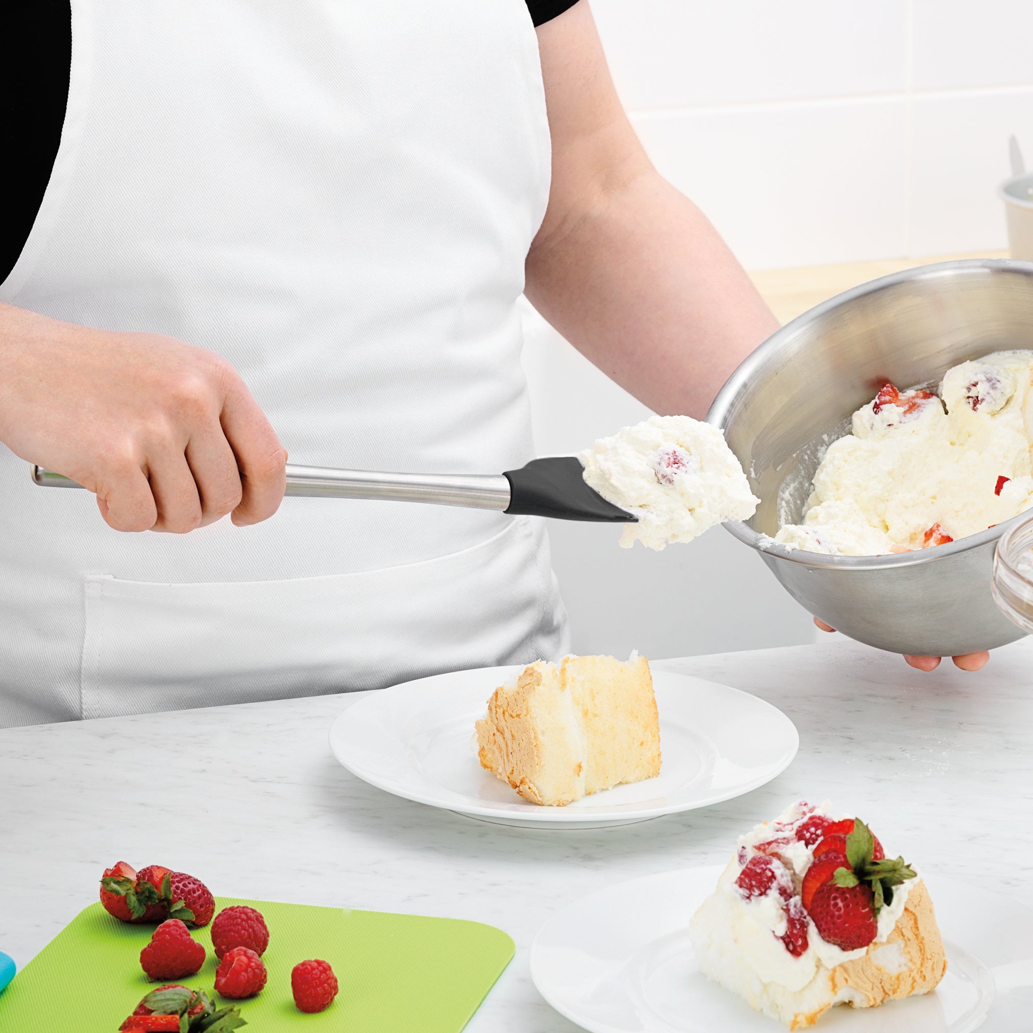Wearing a white apron, a person uses the Flex-Core Spoonula to scoop whipped cream mixed with strawberries onto a cake slice. Fresh strawberries and raspberries are on a green cutting board nearby.
