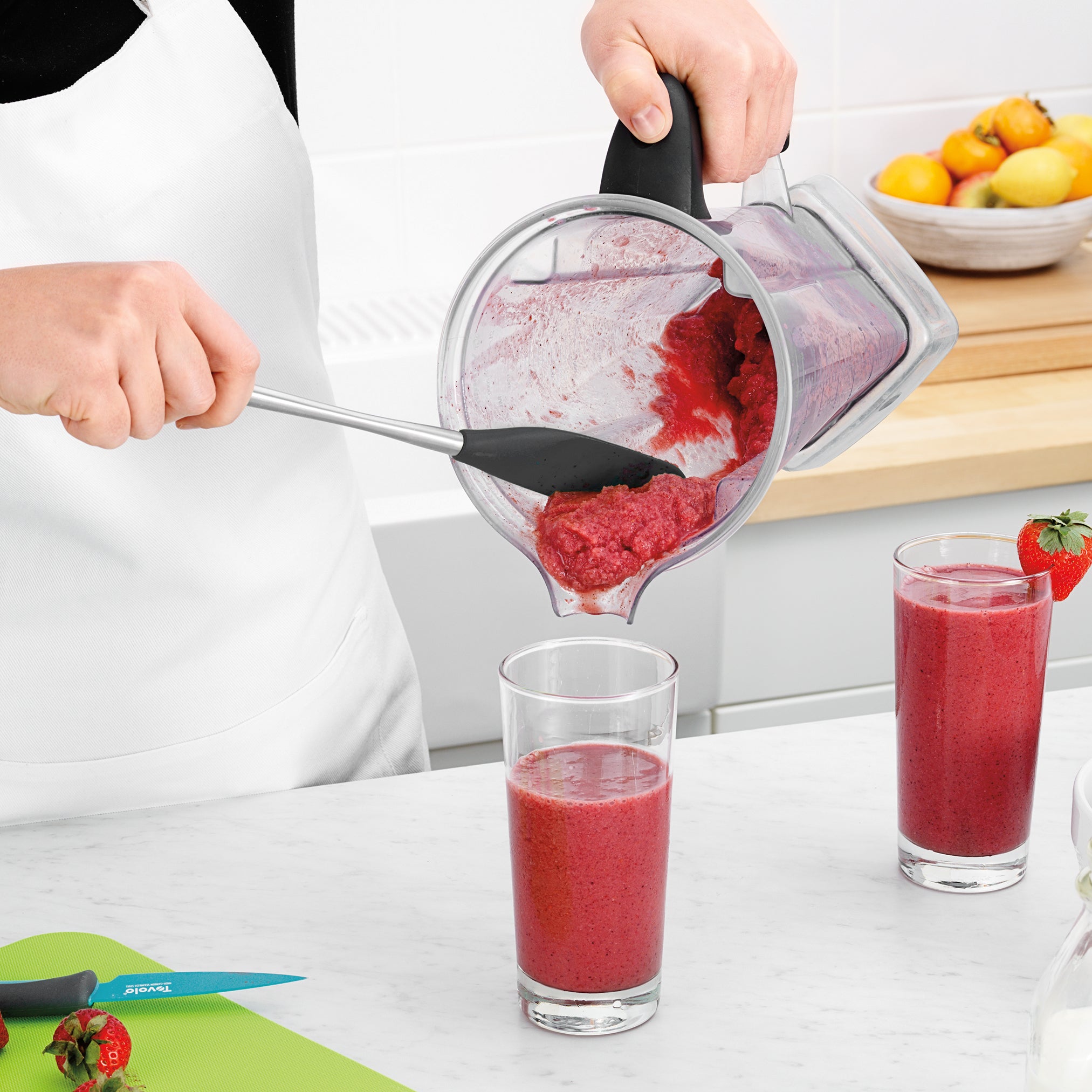 Wearing a white apron, a person uses the Flex-Core Jar Scraper to pour thick red smoothie from a blender into a glass. Another filled glass, strawberries, a blue kitchen tool, and a green cutting board rest on the white counter.