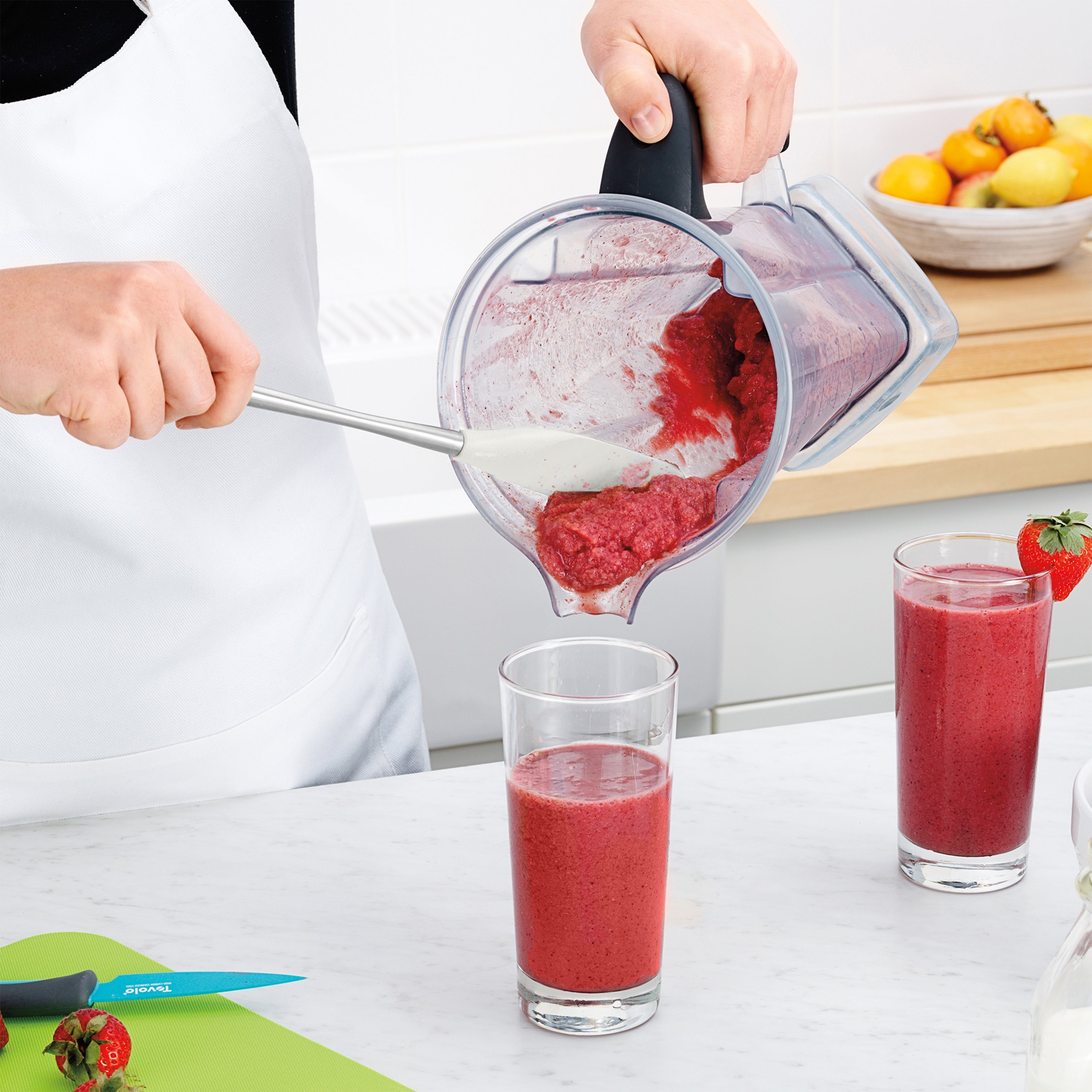 Wearing a white apron, a person uses the Flex-Core Jar Scraper to pour a thick red smoothie from a blender into a glass. On the countertop are another filled glass, a green cutting board, strawberries, and a knife.