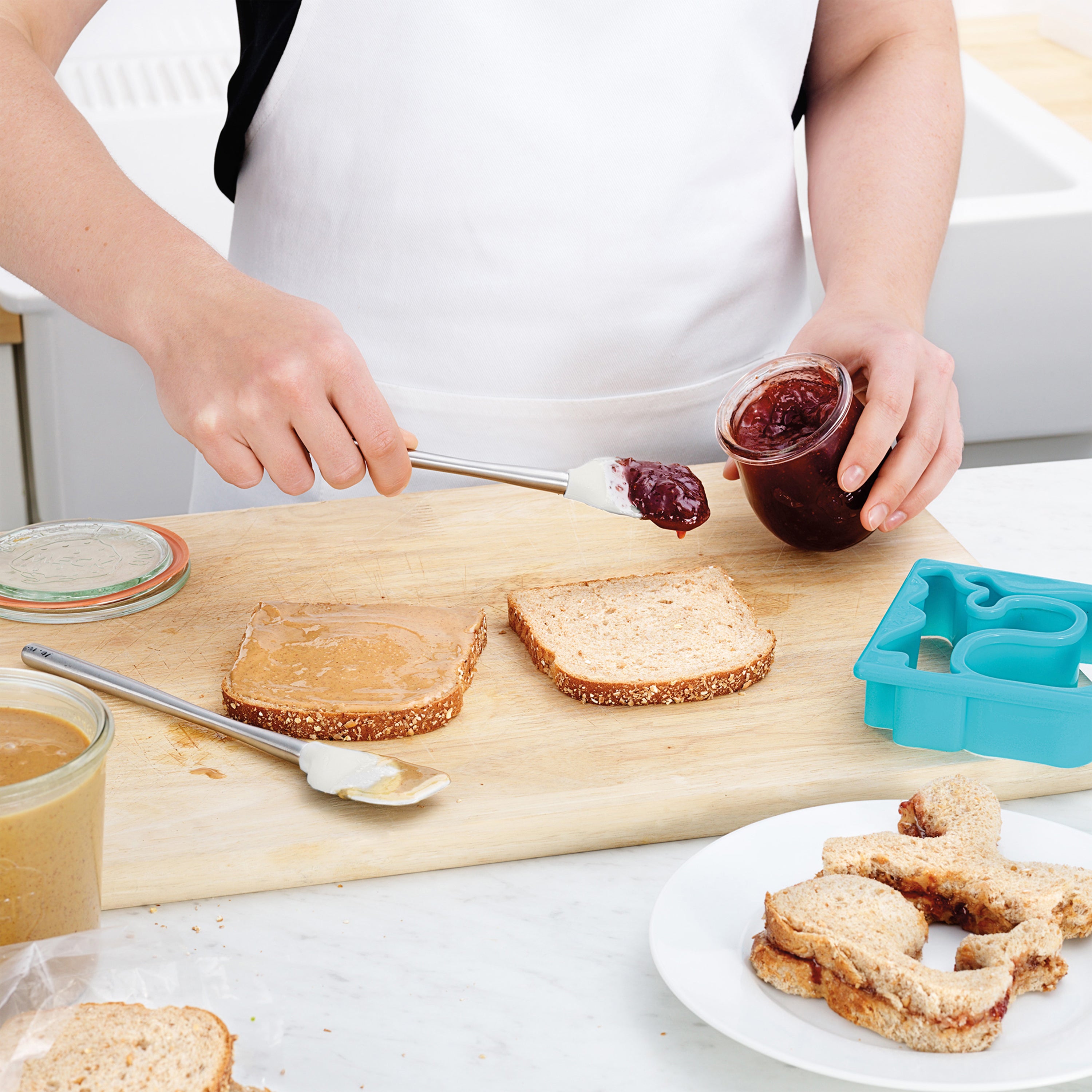 Using the Flex-Core Mini Spat + Spoonula, a person spreads jelly on bread next to a peanut butter slice. A dinosaur-shaped sandwich cutter and finished sandwiches sit on a cutting board.