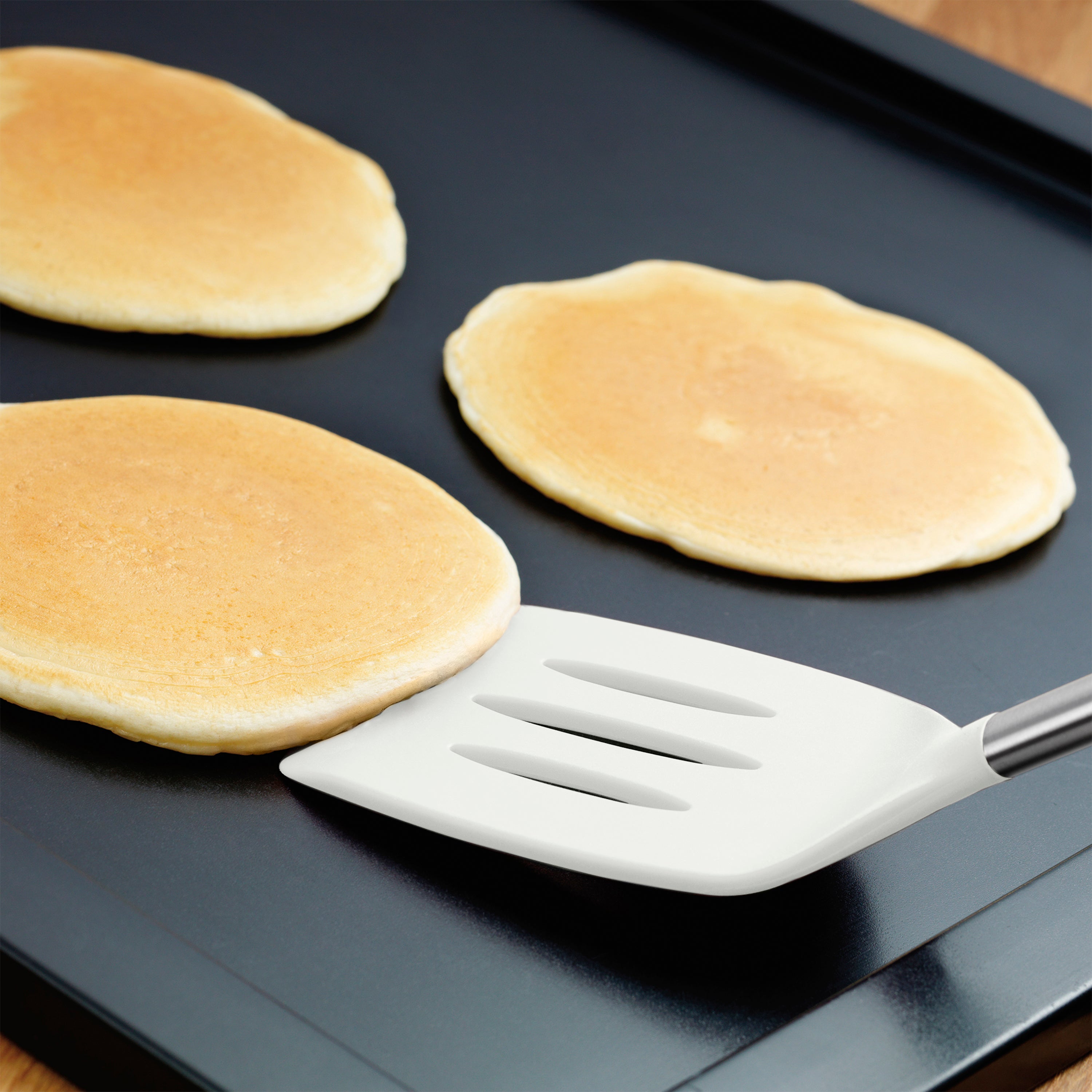 Three golden pancakes cook on a black griddle as a Silicone Slotted Turner, safe for nonstick surfaces, is slid under one pancake, ready to flip it.