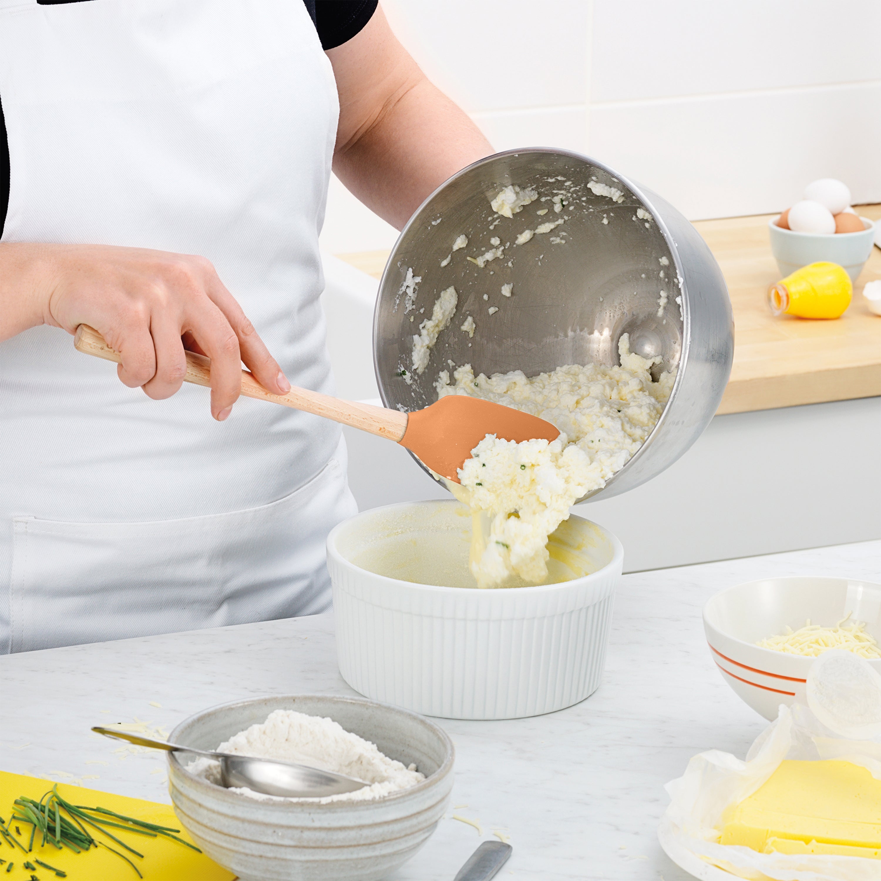 Wearing a white apron, a person uses the Flex-Core Spoonula to transfer a creamy mixture from a metal bowl into a white baking dish, with flour, butter, cheese, and eggs visible on the counter.