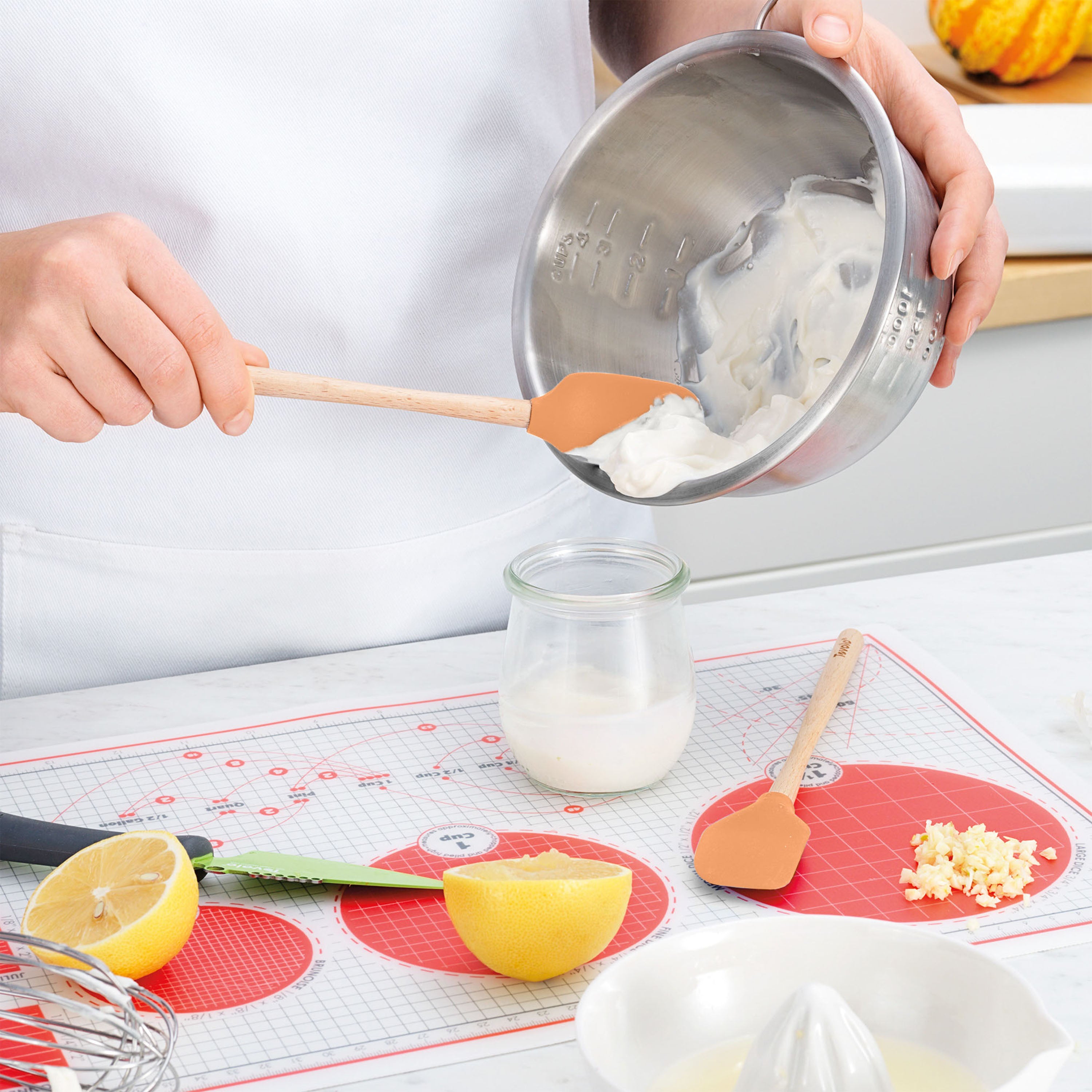 A person uses the Flex-Core Mini Spat + Spoonula to transfer creamy white mixture from a metal bowl into a glass jar on the kitchen counter, with halved lemon, garlic, and utensils nearby.