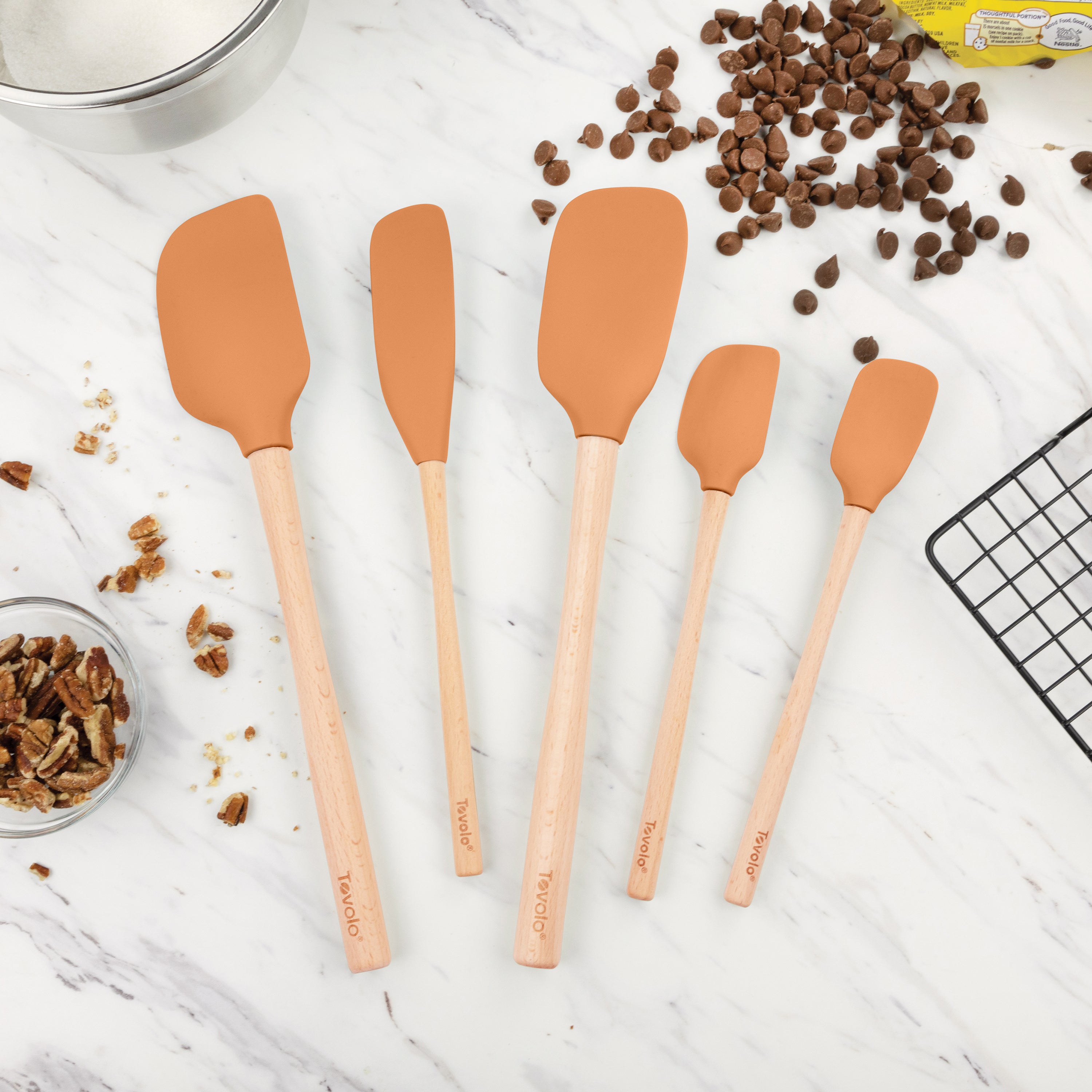 The Flex-Core Wood-Handle Spatula Set of 5 is arranged by size on a white marble countertop, surrounded by chocolate chips, pecans, a wire rack, and baking ingredients—ideal for all your spatula and jar scraper needs.