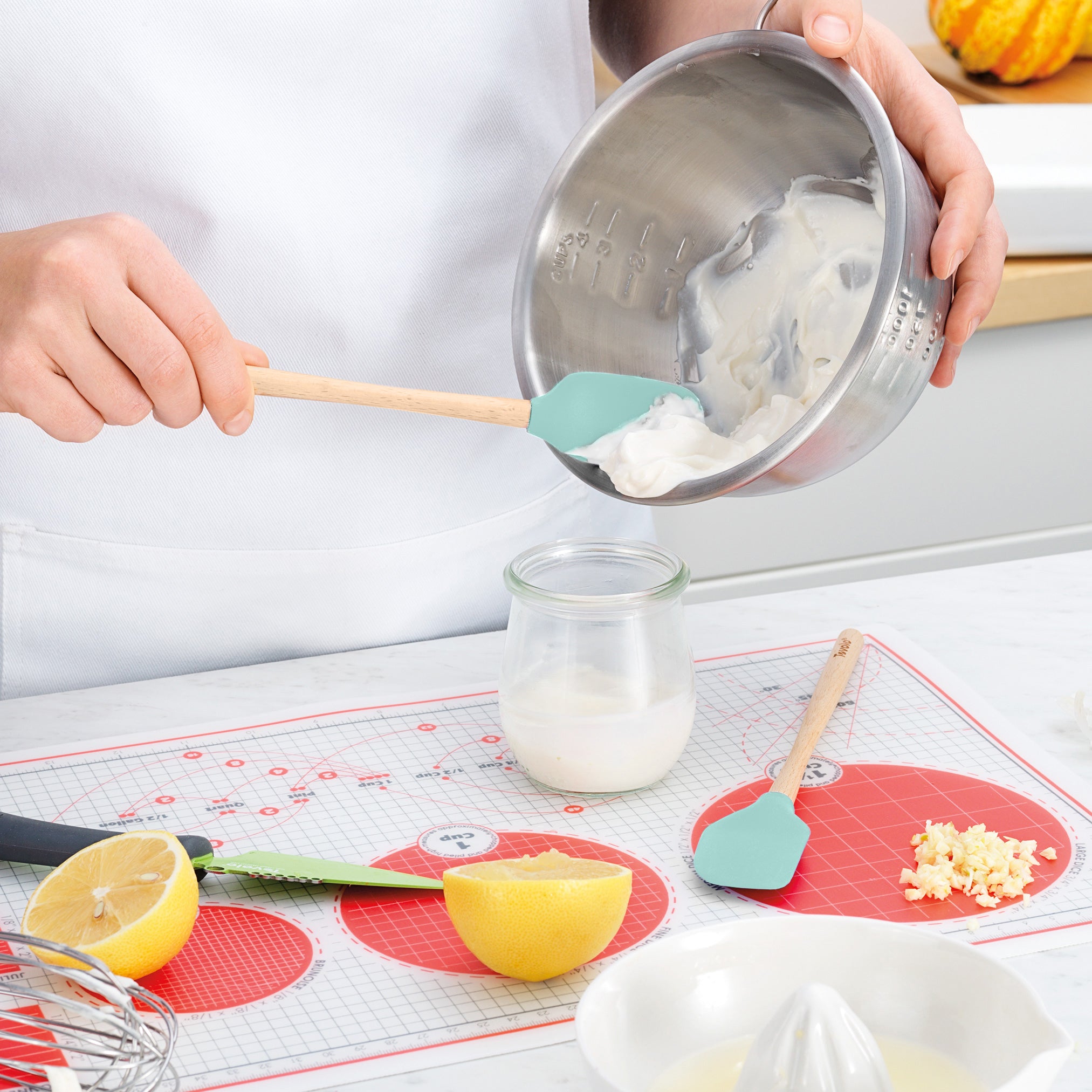 Wearing a white apron, a person uses a Flex-Core Mini Spat + Spoonula to scoop creamy mixture from a metal bowl into a glass jar. Nearby are a halved lemon, knife, minced garlic, and citrus juicer on a red and white mat.
