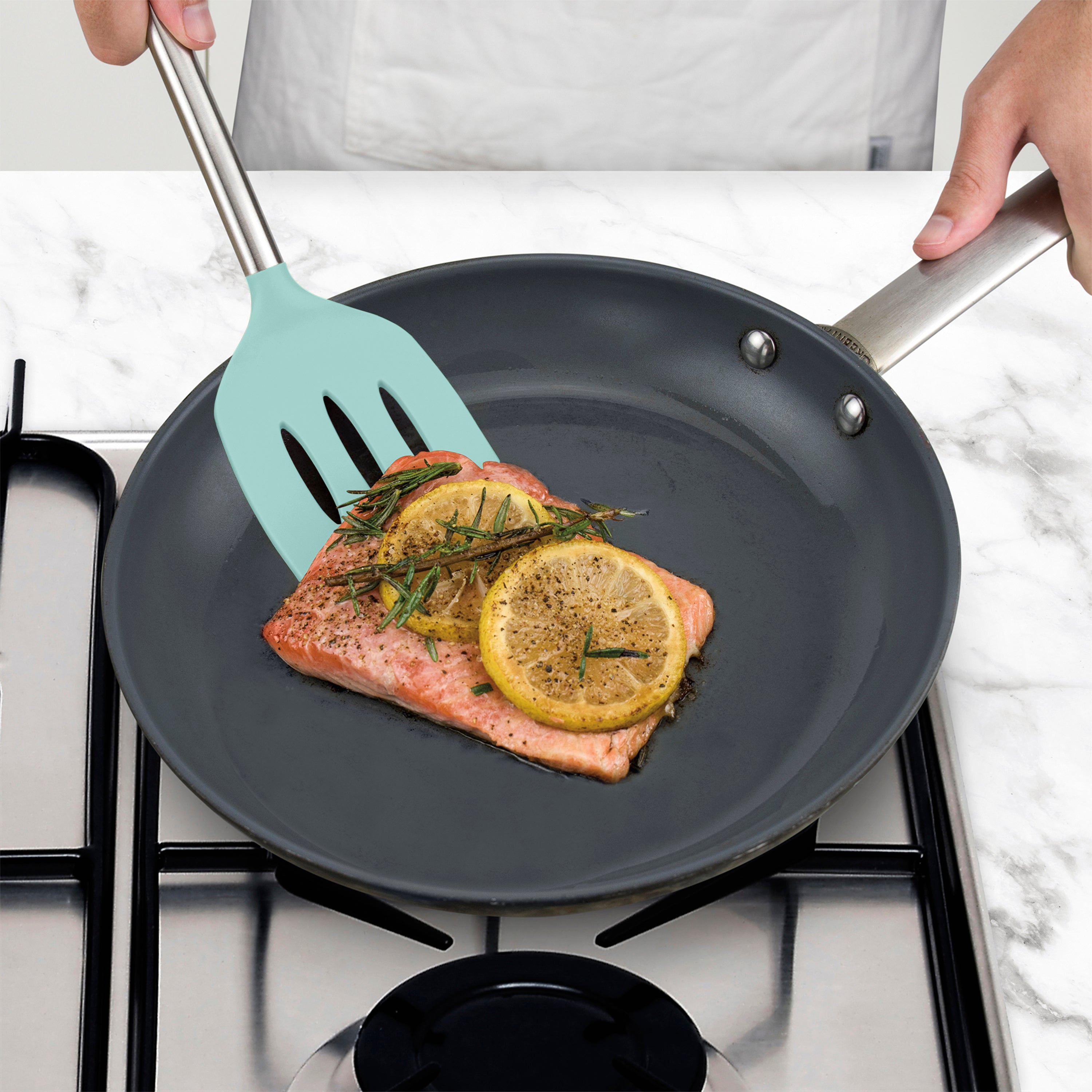 Wearing a white apron, a person uses a Silicone Slotted Turner to cook a salmon fillet with lemon and herbs in a black non-stick frying pan on a gas stove, ensuring the pan’s surface is protected.