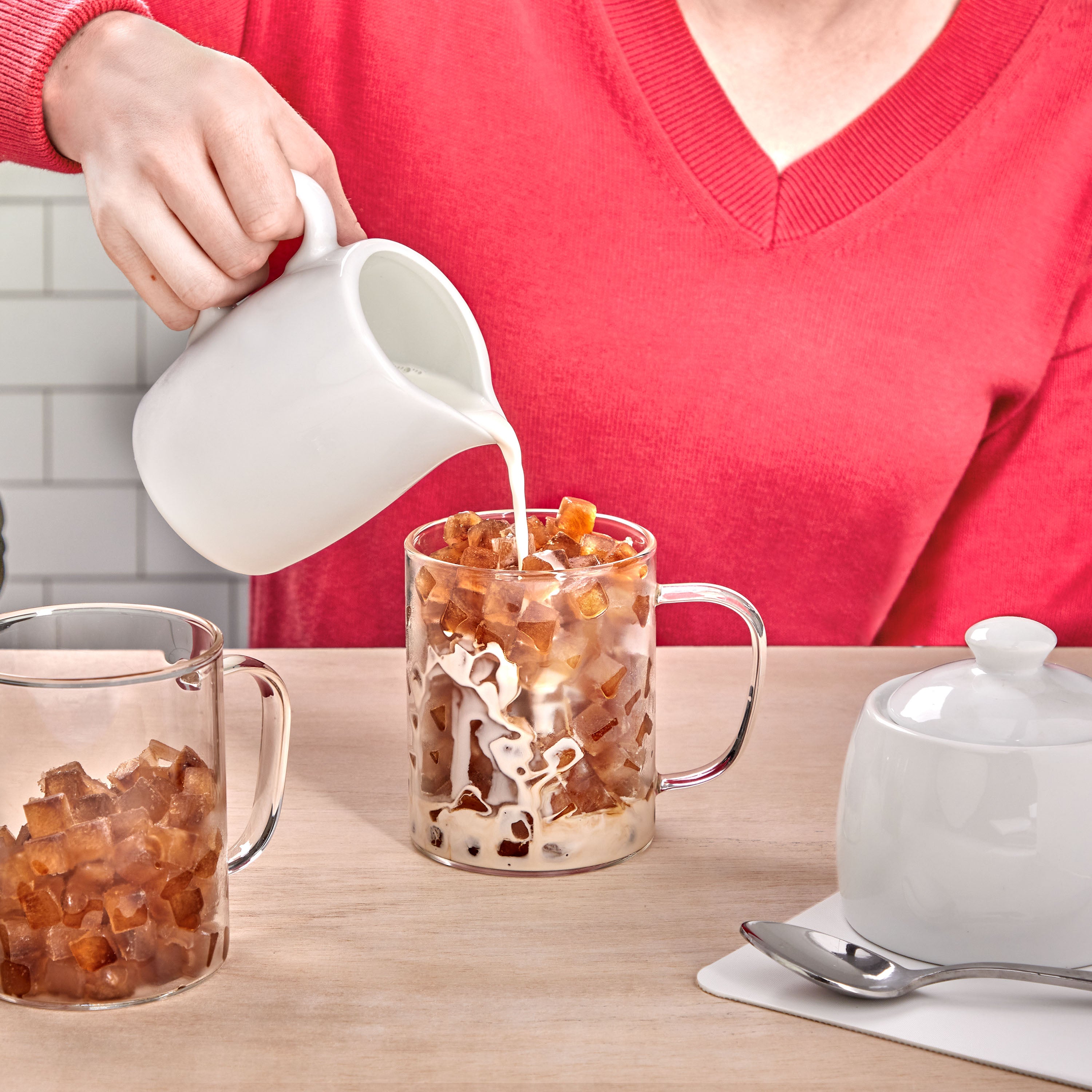 Someone in a red sweater pours milk from a white jug into a glass mug filled with Twist and Release Mini Ice coffee cubes, while another mug and a sugar bowl sit on the table nearby.
