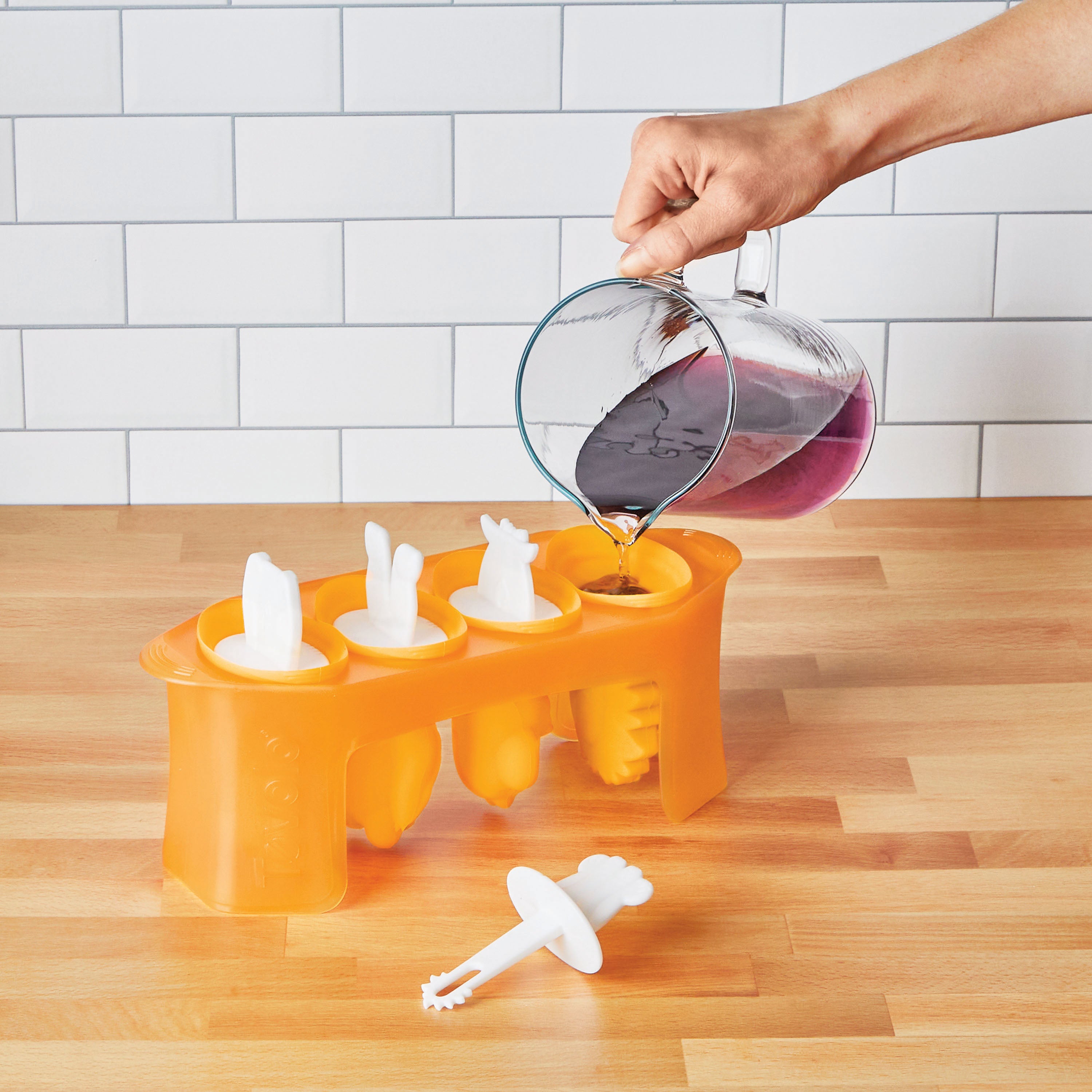 A hand pours purple liquid from a glass measuring cup into orange Pop Molds on a wooden counter to make homemade popsicles. One popsicle stick holder is set aside, with a white tiled wall in the background.
