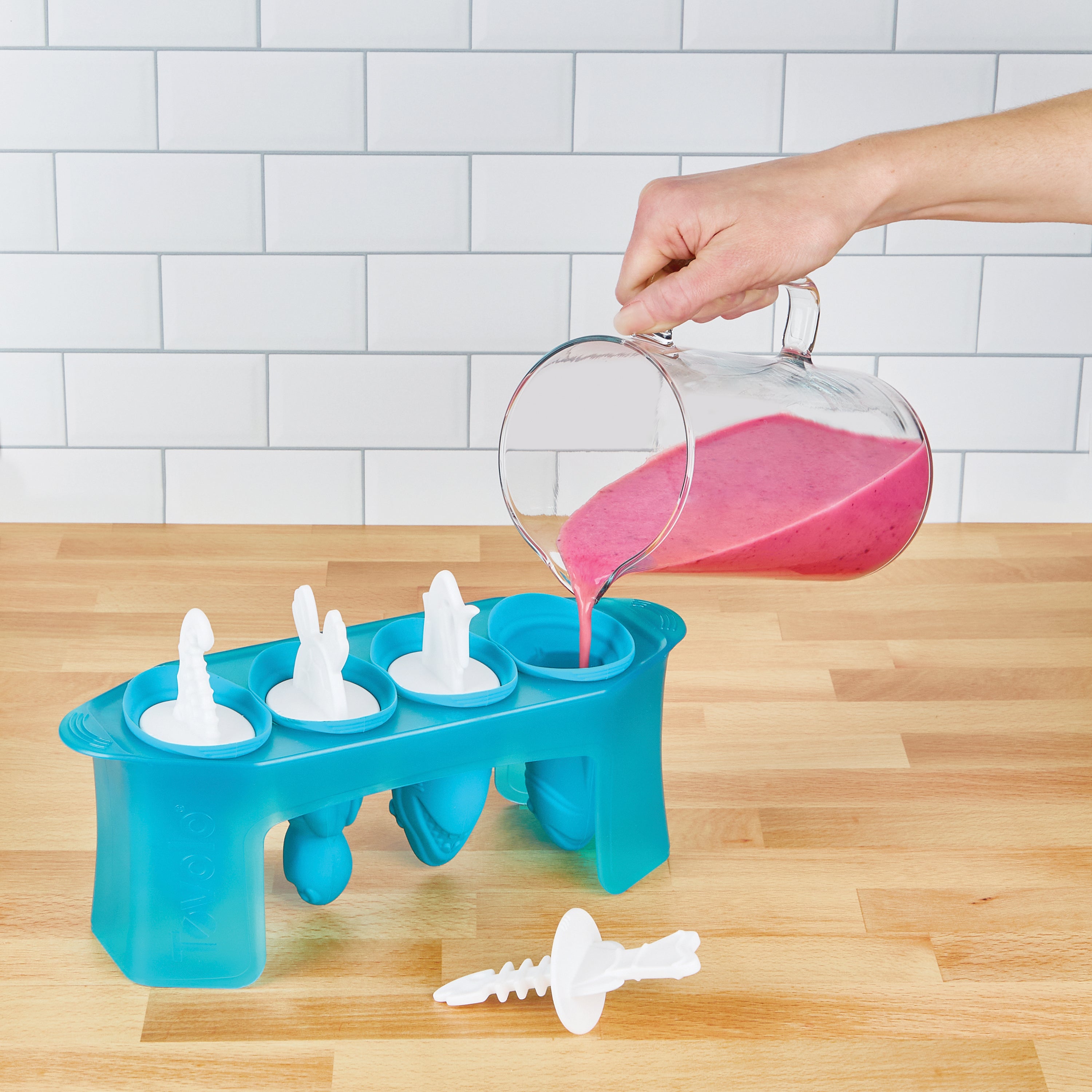 A hand pours pink smoothie from a glass pitcher into Pop Molds—blue popsicle trays with bunny-shaped lids—ideal for making homemade frozen treats, set on a wooden countertop before a white tiled wall.
