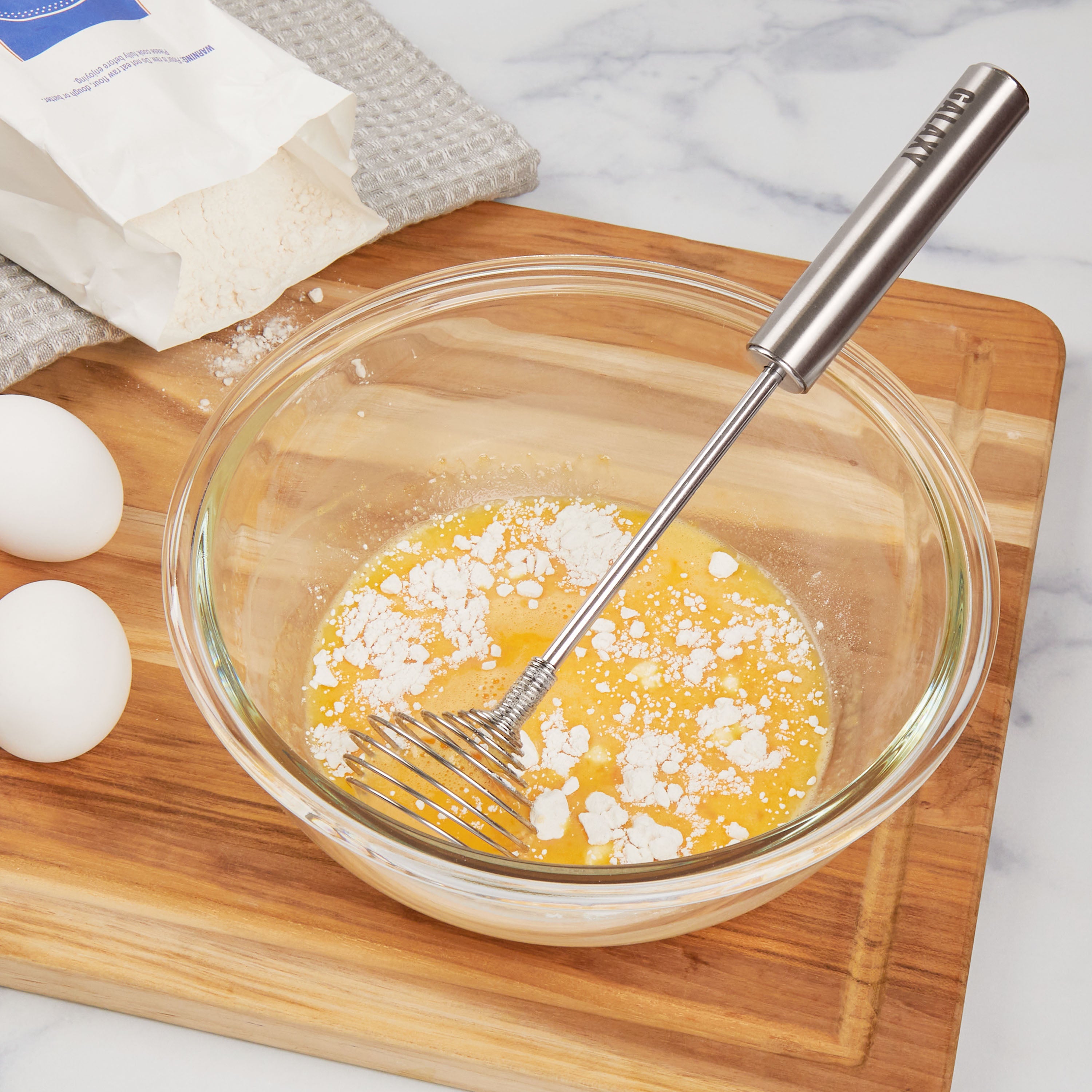 A glass bowl with eggs and flour being mixed using a Specialty Whisk sits on a wooden cutting board, with two eggs and a bag of flour nearby on the counter.