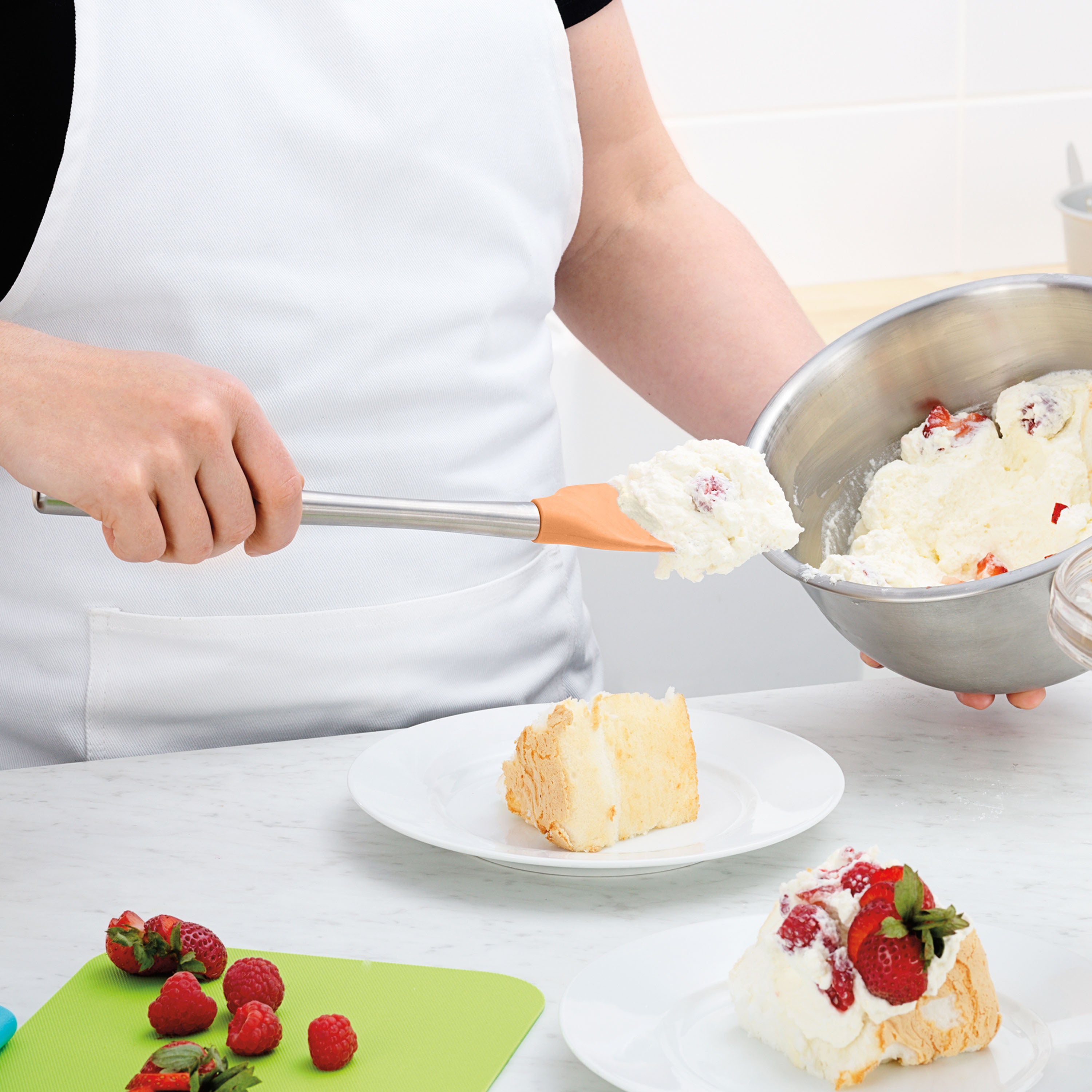 Wearing a white apron, a person uses the Flex-Core Spoonula to add whipped cream and strawberries onto a cake slice on a plate. Fresh strawberries and a green cutting board rest on the marble counter nearby.