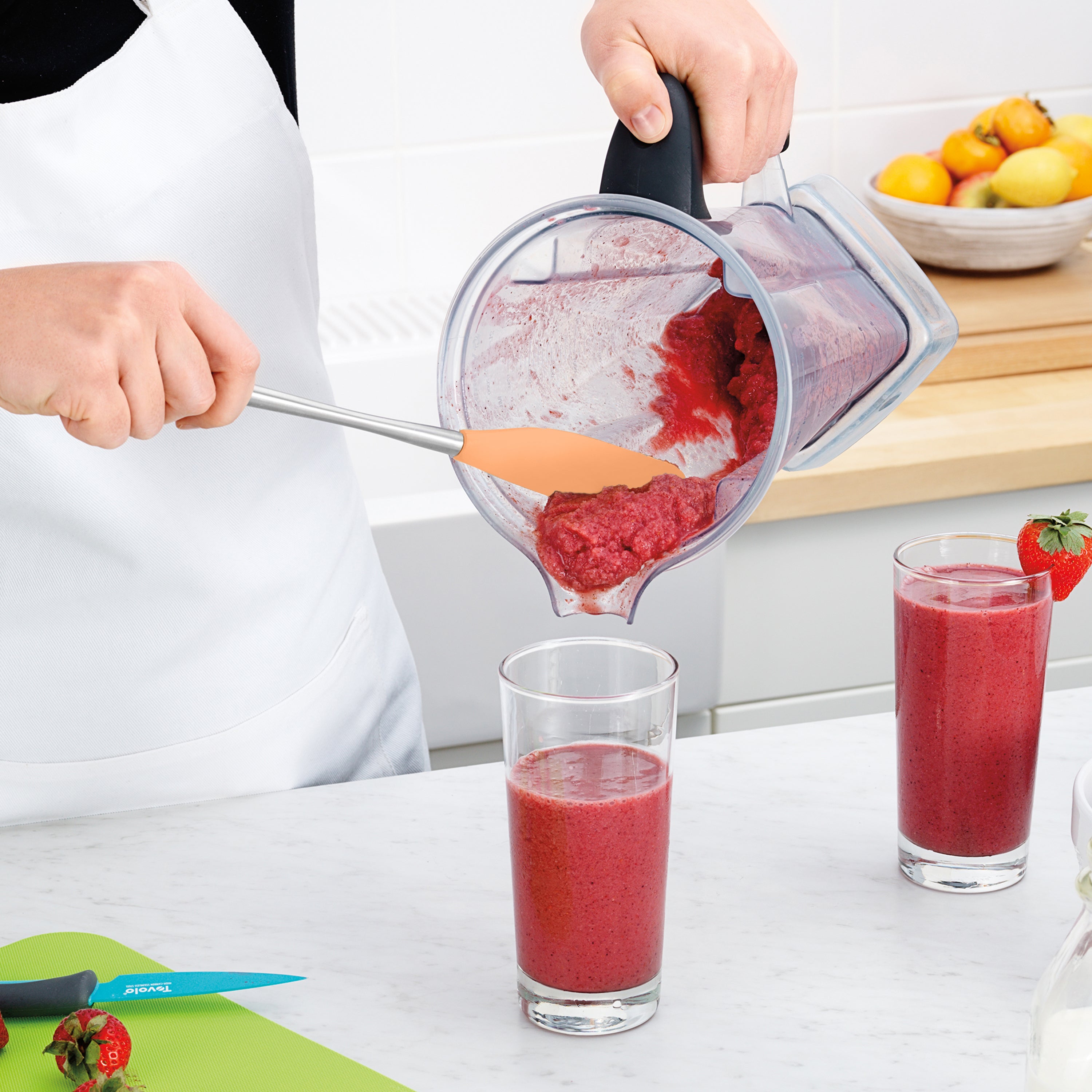 Wearing a white apron, a person uses the Flex-Core Jar Scraper to pour thick red smoothie from a blender into a glass. On the kitchen counter are two filled glasses, strawberries, a blue knife, and a bowl of fruit.