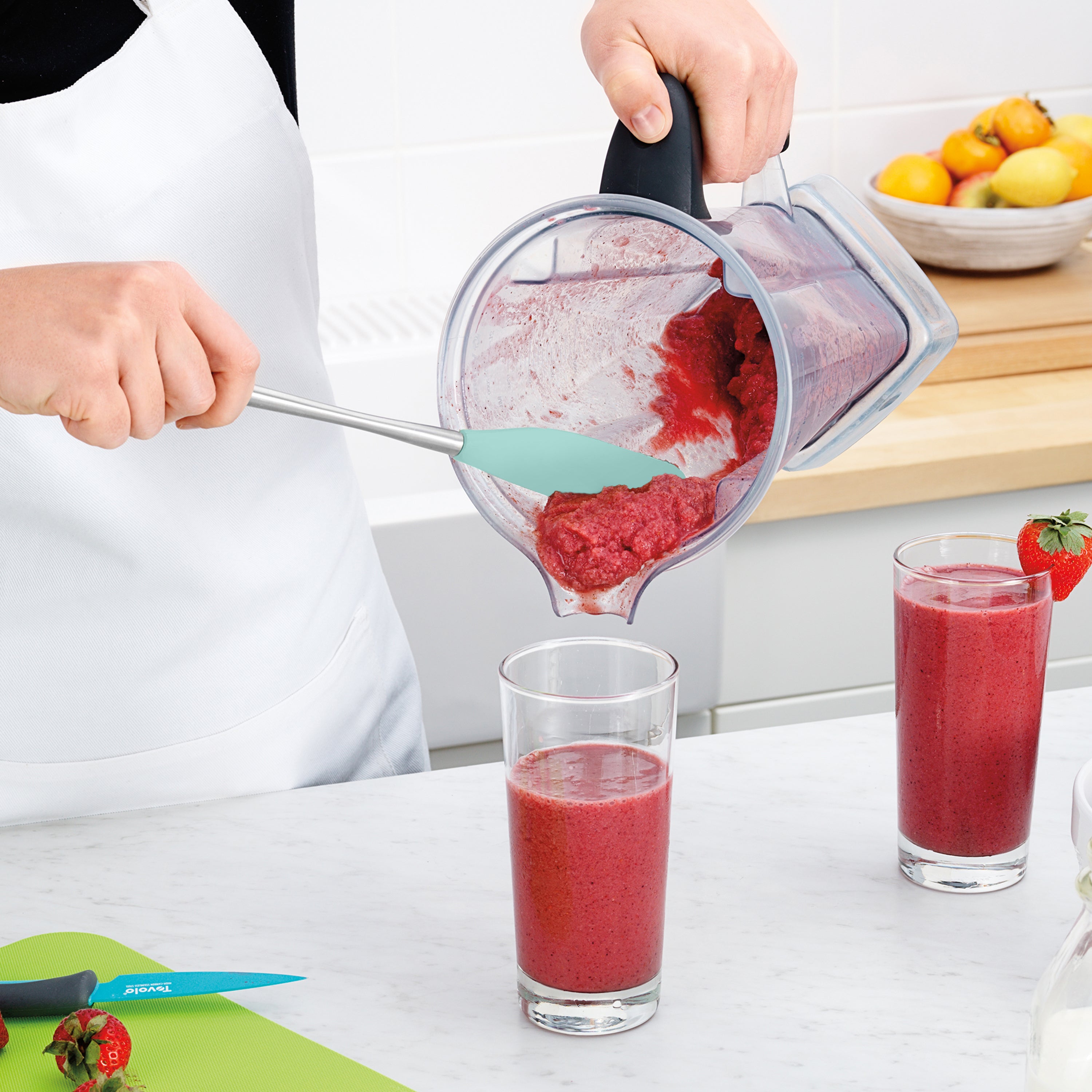 Wearing a white apron, a person uses the Flex-Core Jar Scraper to pour a thick red smoothie from a blender into a glass on the kitchen counter, beside another filled glass, a knife, and strawberries.