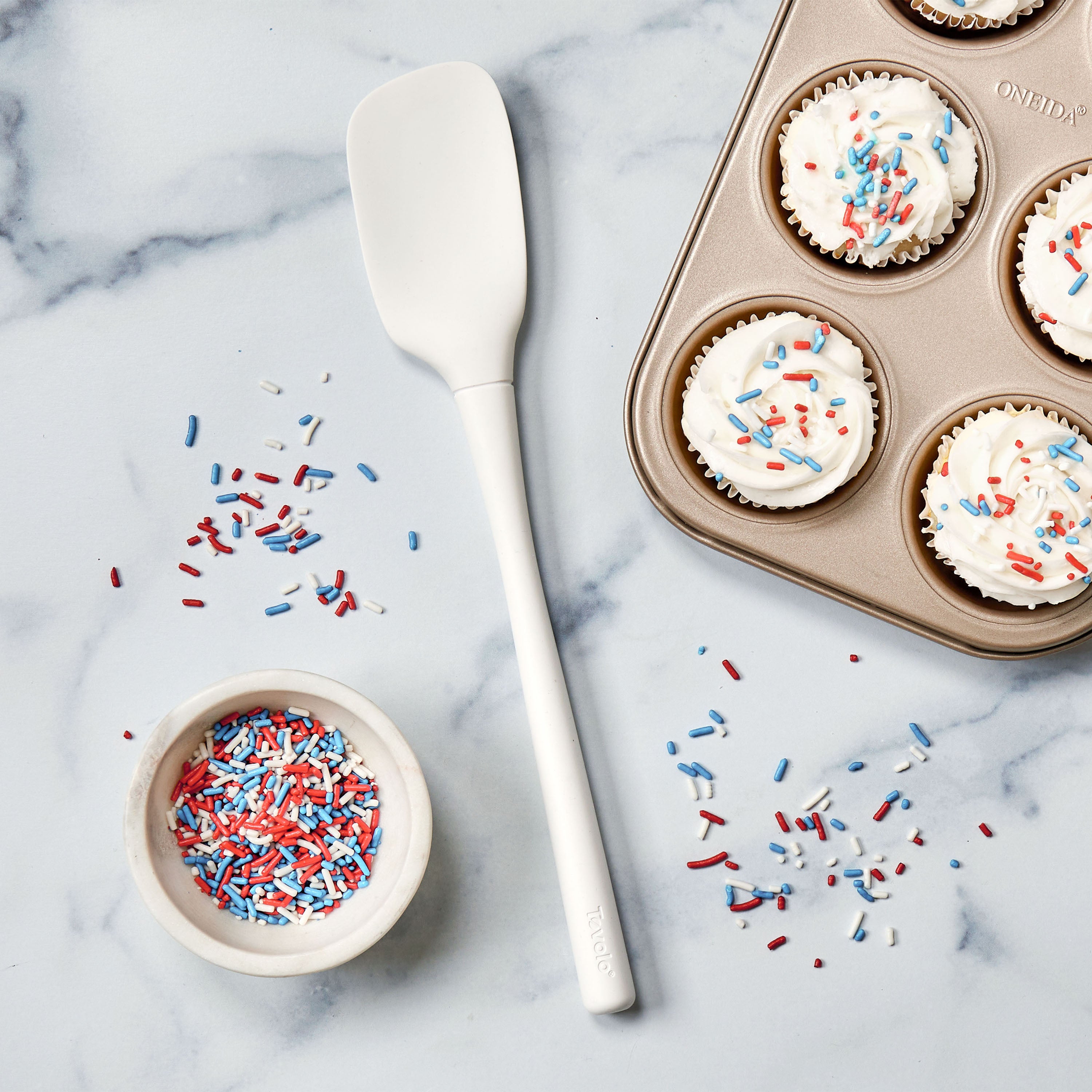A white Flex-Core Spoonula, a bowl of red, white, and blue sprinkles, and a muffin tray with frosted cupcakes topped with sprinkles are displayed on a marble surface.
