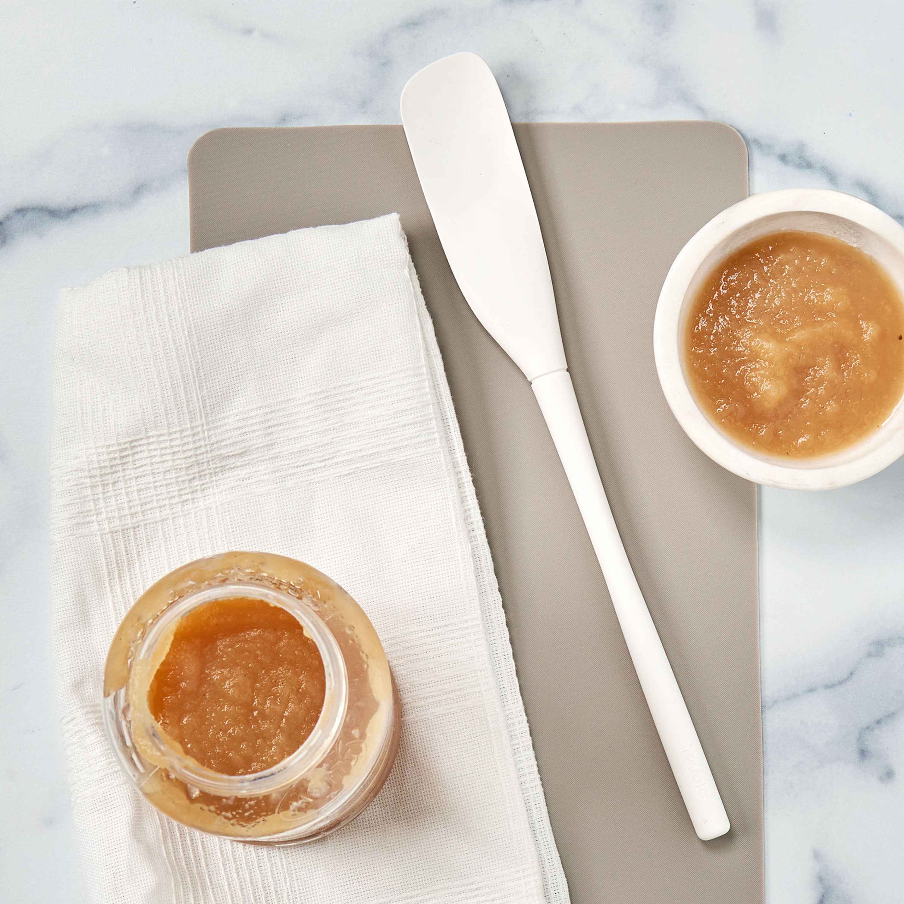 A jar and a small bowl of light brown applesauce sit on a napkin and gray cutting board, with the white Flex-Core Jar Scraper nearby, all displayed on a marble surface.