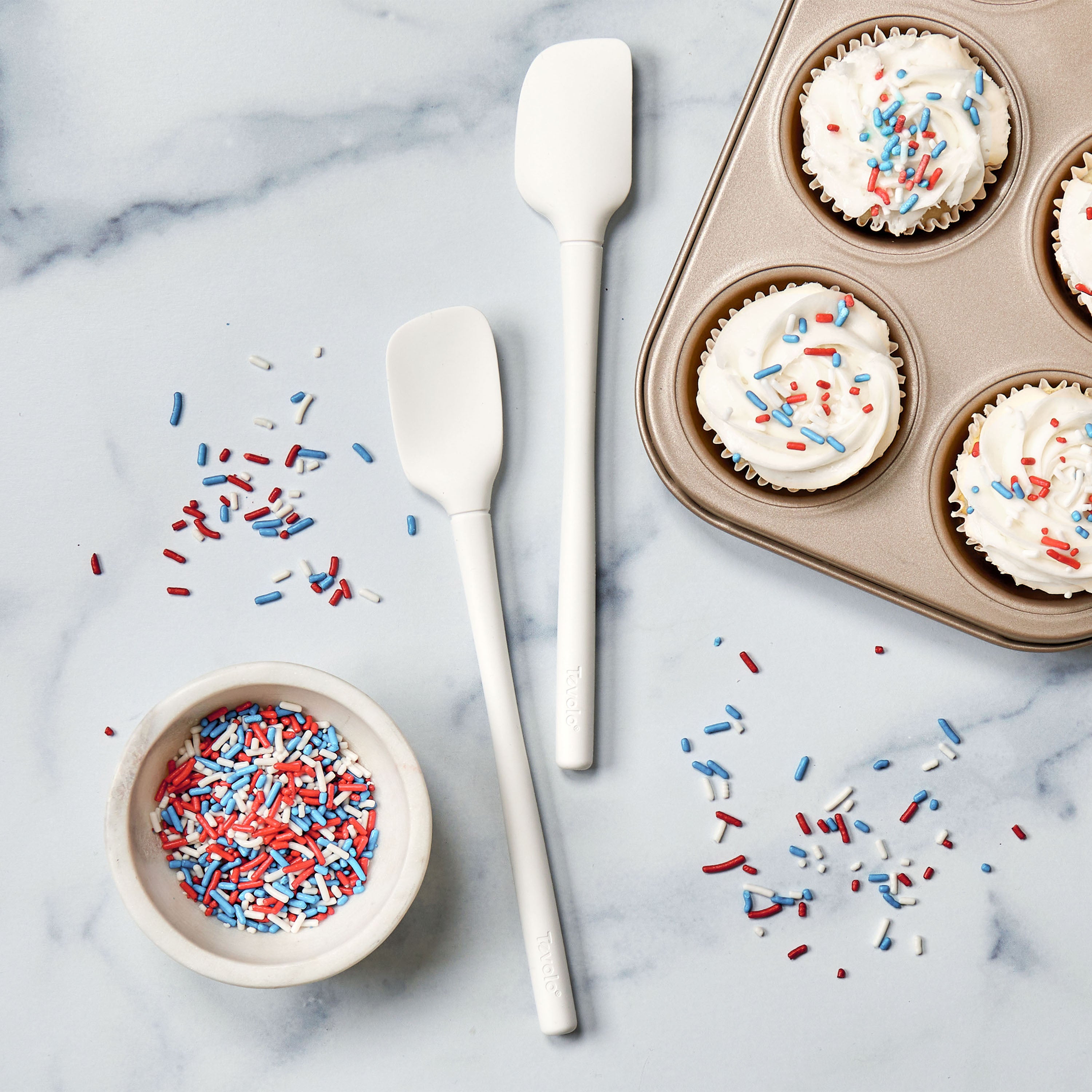 A bowl of red, white, and blue sprinkles sits on marble next to a Flex-Core Mini Spat + Spoonula and a muffin tin filled with cupcakes topped with white frosting and sprinkles, with some sprinkles scattered nearby.