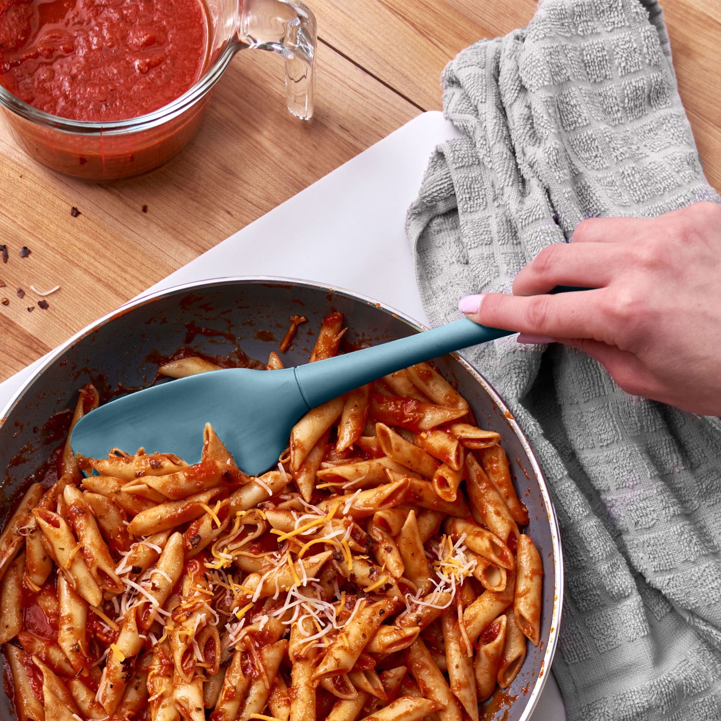 A hand stirs penne pasta with tomato sauce and shredded cheese in a pan using a Flex-Core Spoonula, beside a gray towel and a glass measuring cup of sauce on a wooden surface.