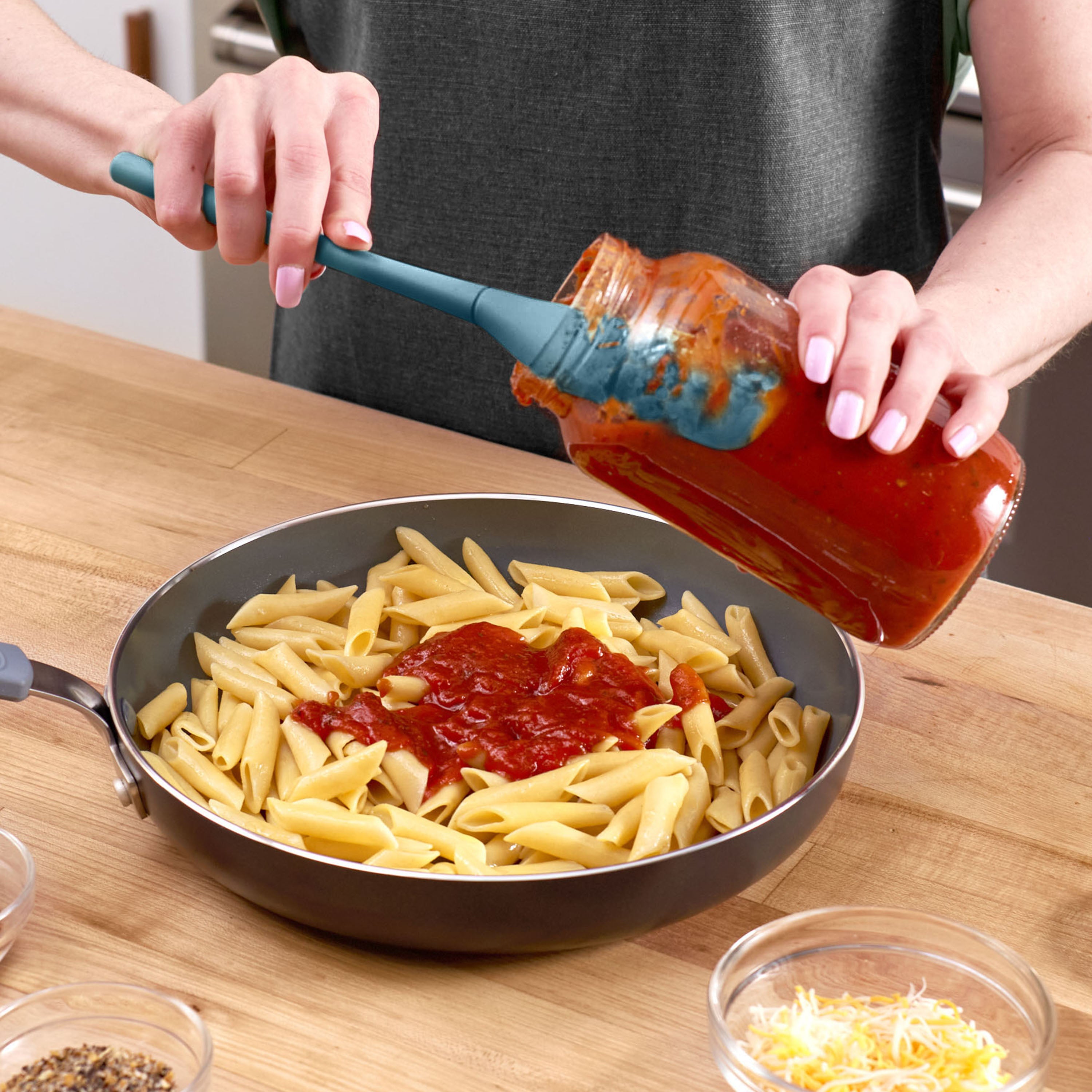 A person uses the Flex-Core Jar Scraper to pour red pasta sauce from a jar onto cooked penne pasta in a skillet, preparing a meal on a wooden countertop with small bowls of ingredients nearby.