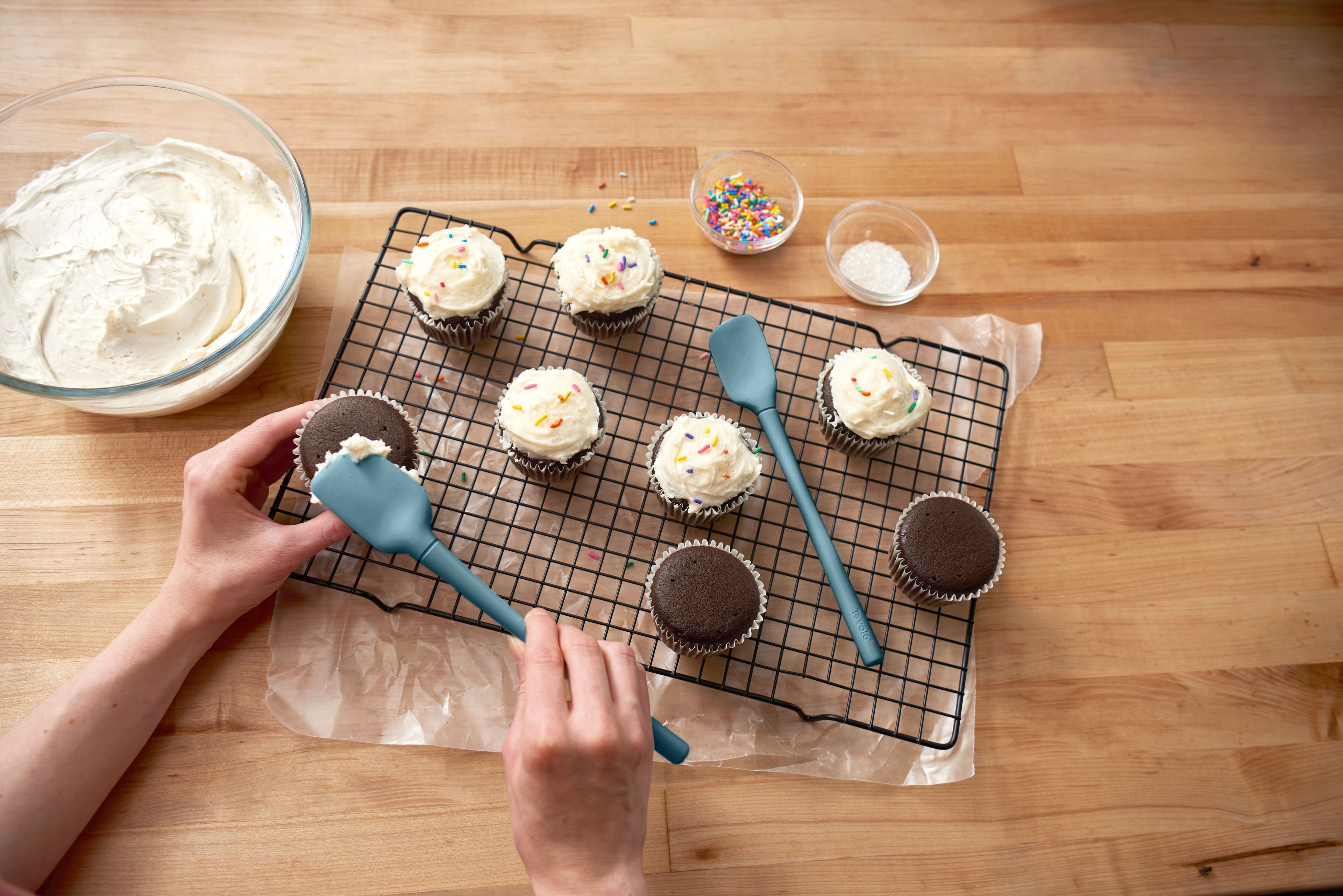 Using the Flex-Core Mini Spat + Spoonula, someone decorates chocolate cupcakes with white frosting and sprinkles. The cupcakes rest on a cooling rack, surrounded by bowls of frosting, sprinkles, and sugar on a wooden surface.