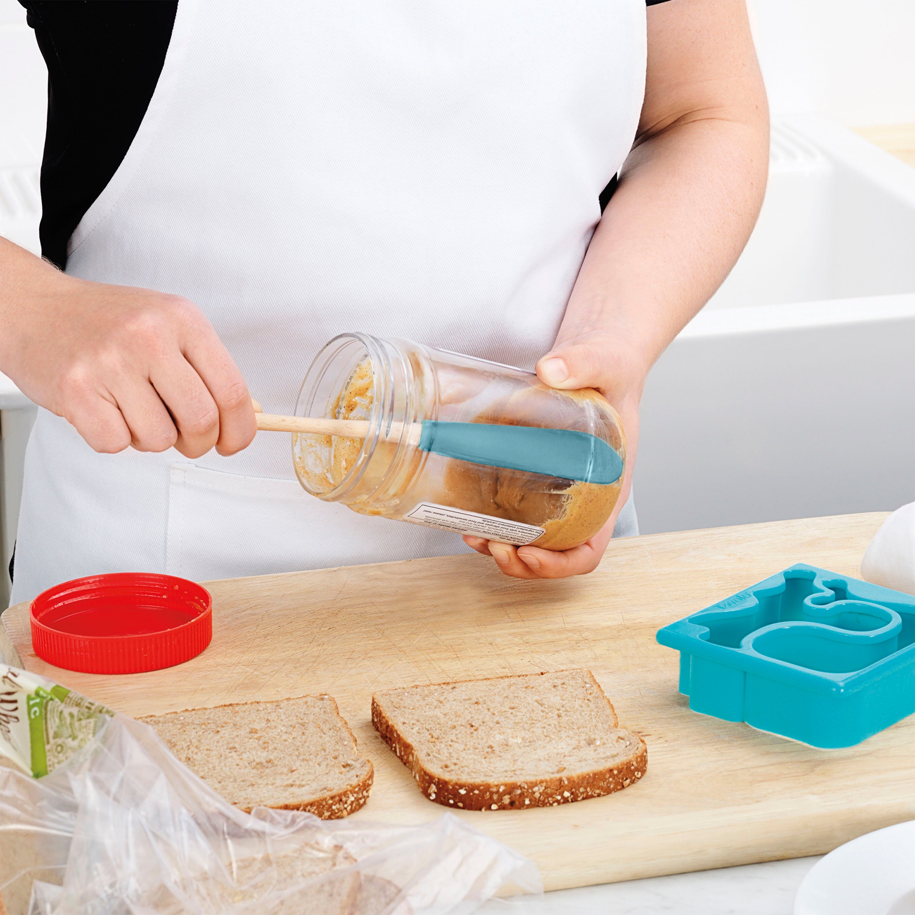 Wearing a white apron, a person uses the Flex-Core Jar Scraper to spread peanut butter onto whole wheat sandwich bread, with a sandwich cutter, plastic bread bag, and other kitchen tools nearby on a wooden surface.