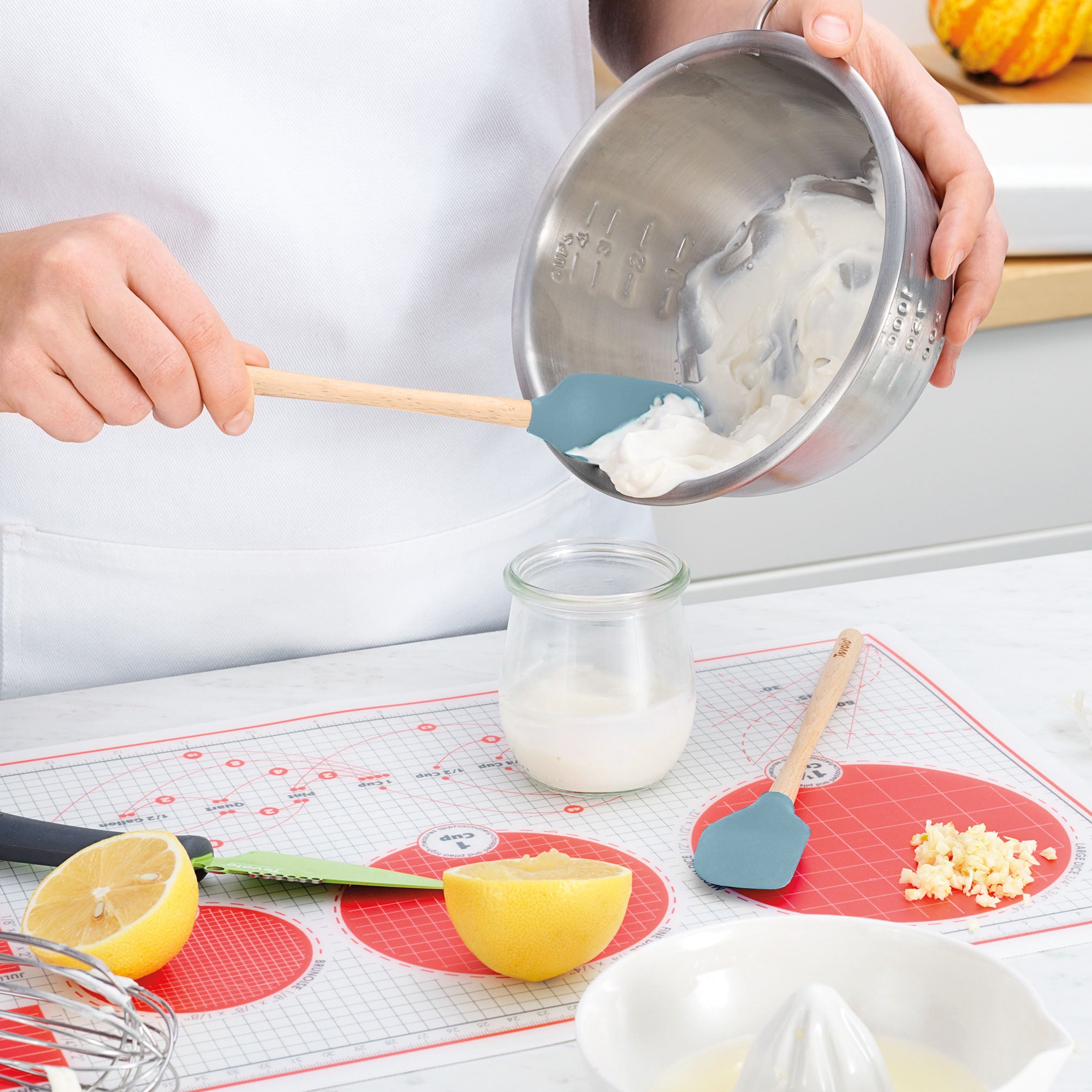 A person uses a blue Flex-Core Mini Spat + Spoonula to transfer creamy white mixture from a metal bowl to a glass jar. On the counter are a halved lemon, a knife, minced garlic, another spatula, and a whisk.