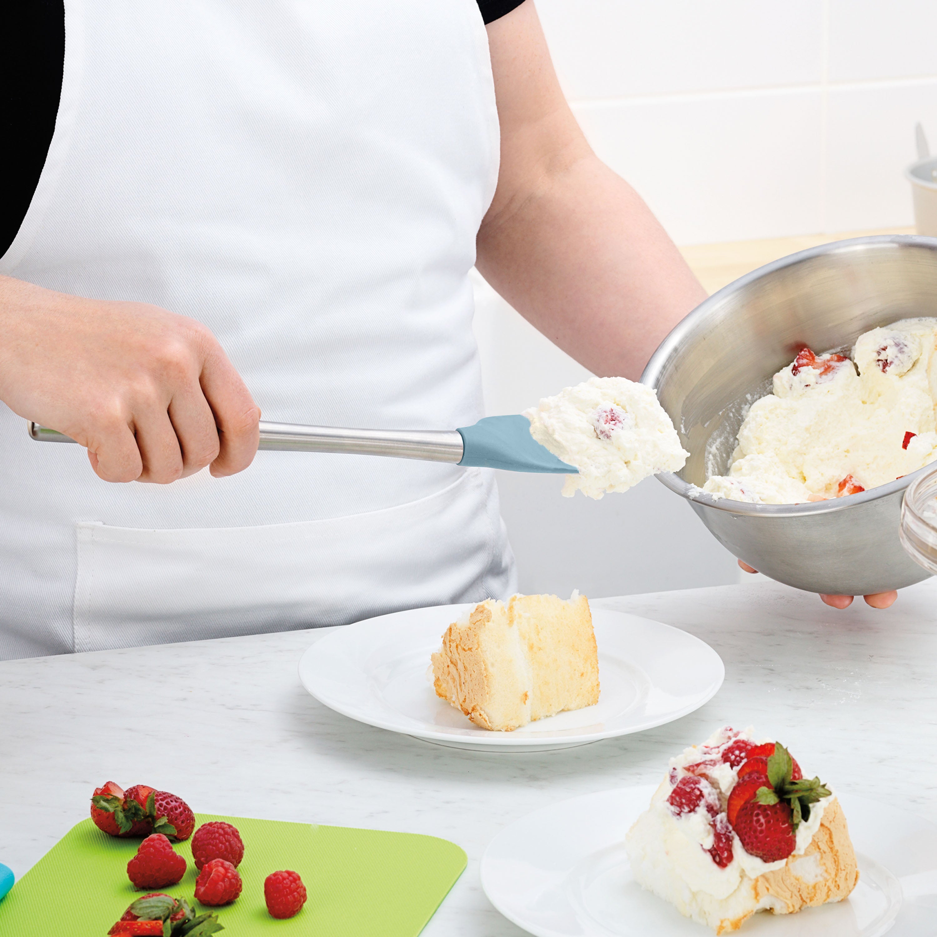 Wearing a white apron, a person uses the Flex-Core Spoonula to add whipped cream with strawberries to a cake slice on a plate, while fresh strawberries and raspberries rest on a nearby cutting board.