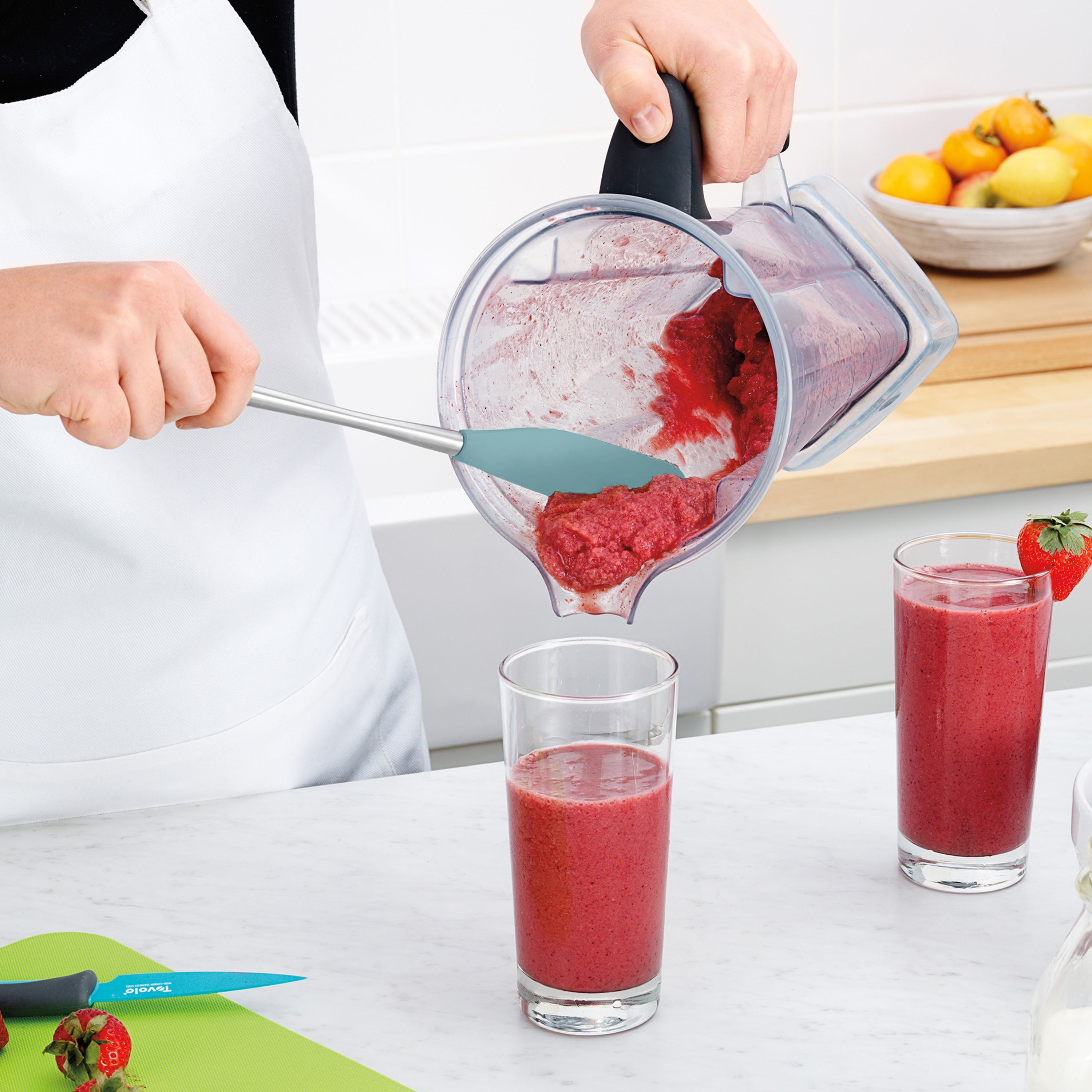Wearing a white apron, a person uses the Flex-Core Jar Scraper to pour a thick red smoothie from a blender into a glass on the kitchen counter, surrounded by another full glass, a knife, strawberries, and a bowl of fruit.
