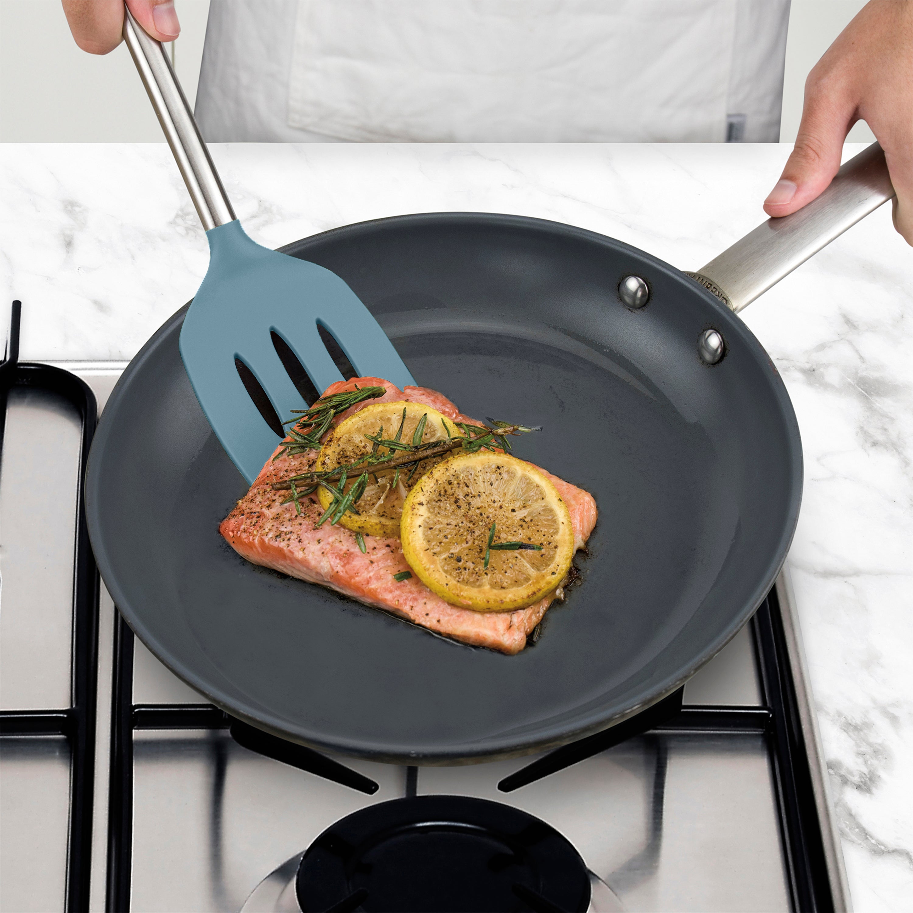 Using the Silicone Slotted Turner, a person cooks a piece of salmon with lemon slices and herbs in a frying pan on the stovetop, ensuring the nonstick surface stays safe.