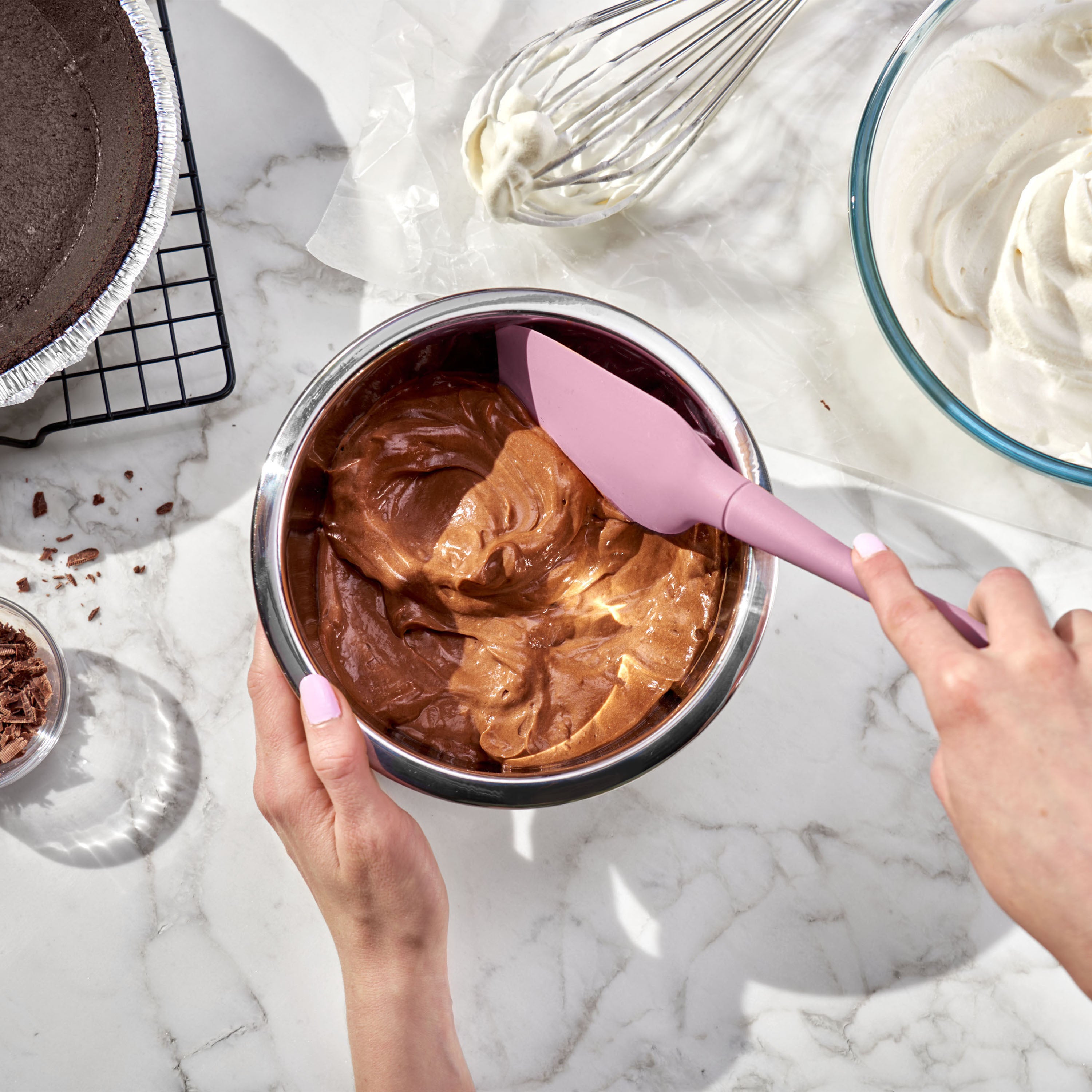 Someone mixes chocolate mousse in a metal bowl with a pink Flex-Core Spatula on a marble countertop, surrounded by chocolate pie crust, whipped cream, a whisk, and chocolate shavings.