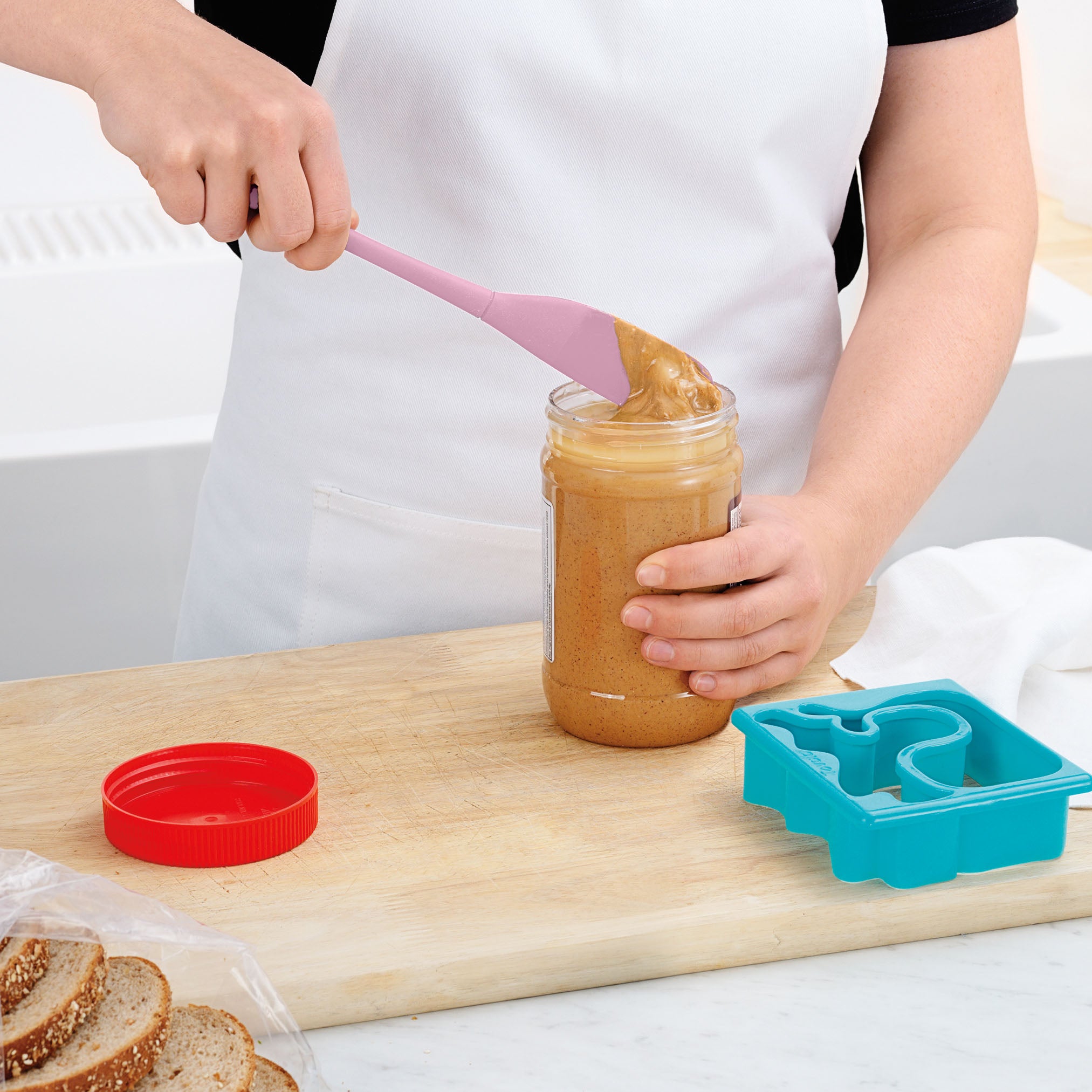 A person in a white apron uses the Flex-Core Jar Scraper, a pink silicone spatula, to scoop peanut butter from a jar on the kitchen counter. Sliced bread, a red lid, and a blue sandwich cutter are also visible.