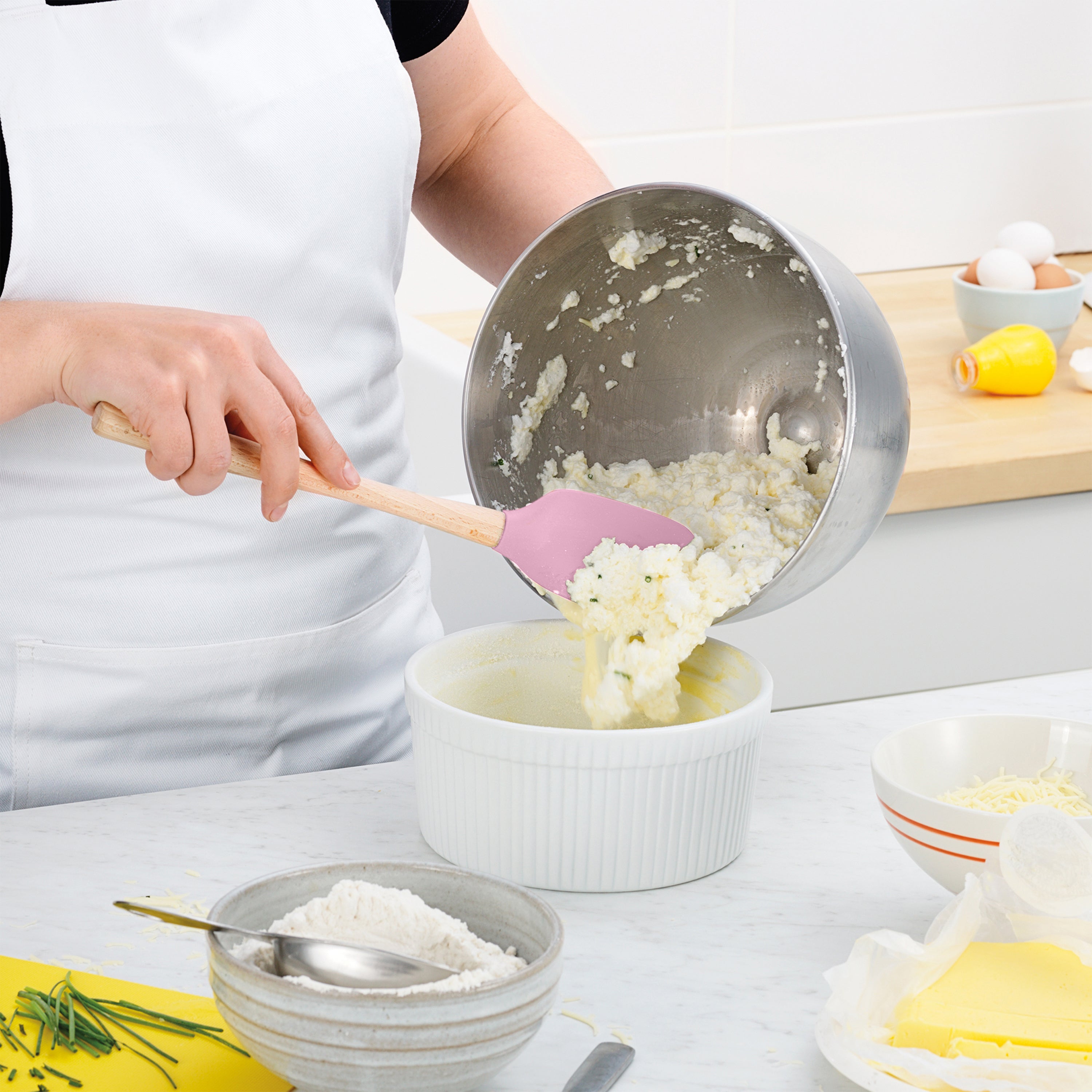 Wearing a white apron, a person uses a Flex-Core Spoonula to scoop creamy mixture from a metal bowl into a white baking dish, with assorted baking ingredients neatly placed on the kitchen counter.