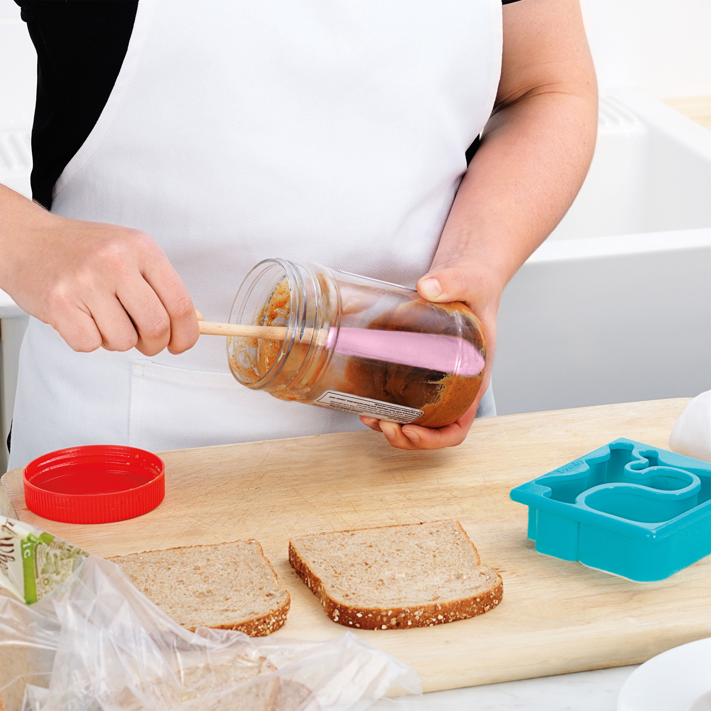 A person in a white apron spreads peanut butter from a jar onto bread using a Flex-Core Jar Scraper. Two slices of bread and a blue sandwich cutter sit on the wooden counter.