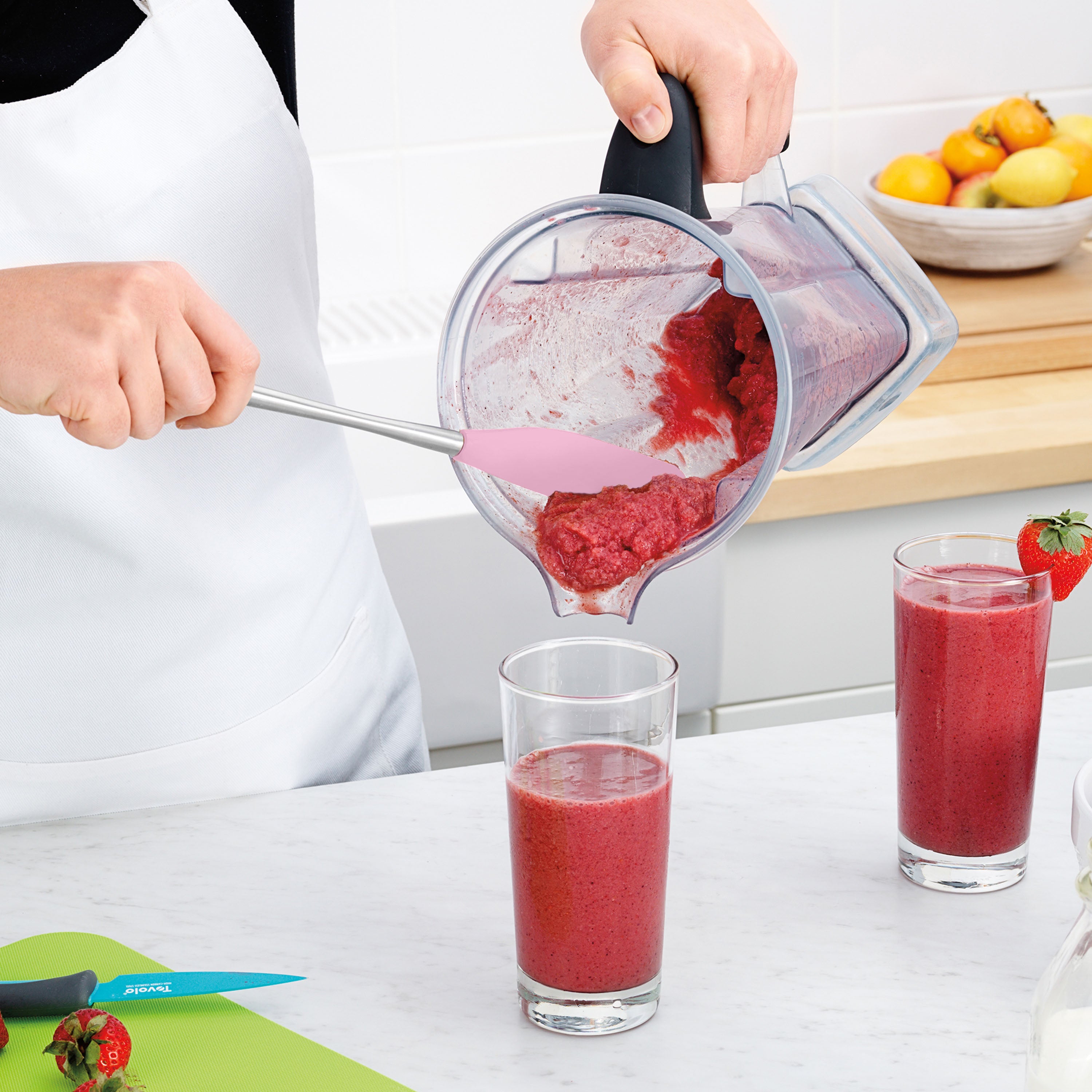 Wearing a white apron, a person uses the Flex-Core Jar Scraper to pour a thick red smoothie from a blender into glasses. On the counter: two filled glasses, fresh strawberries on a green cutting board, and the Flex-Core Jar Scraper.