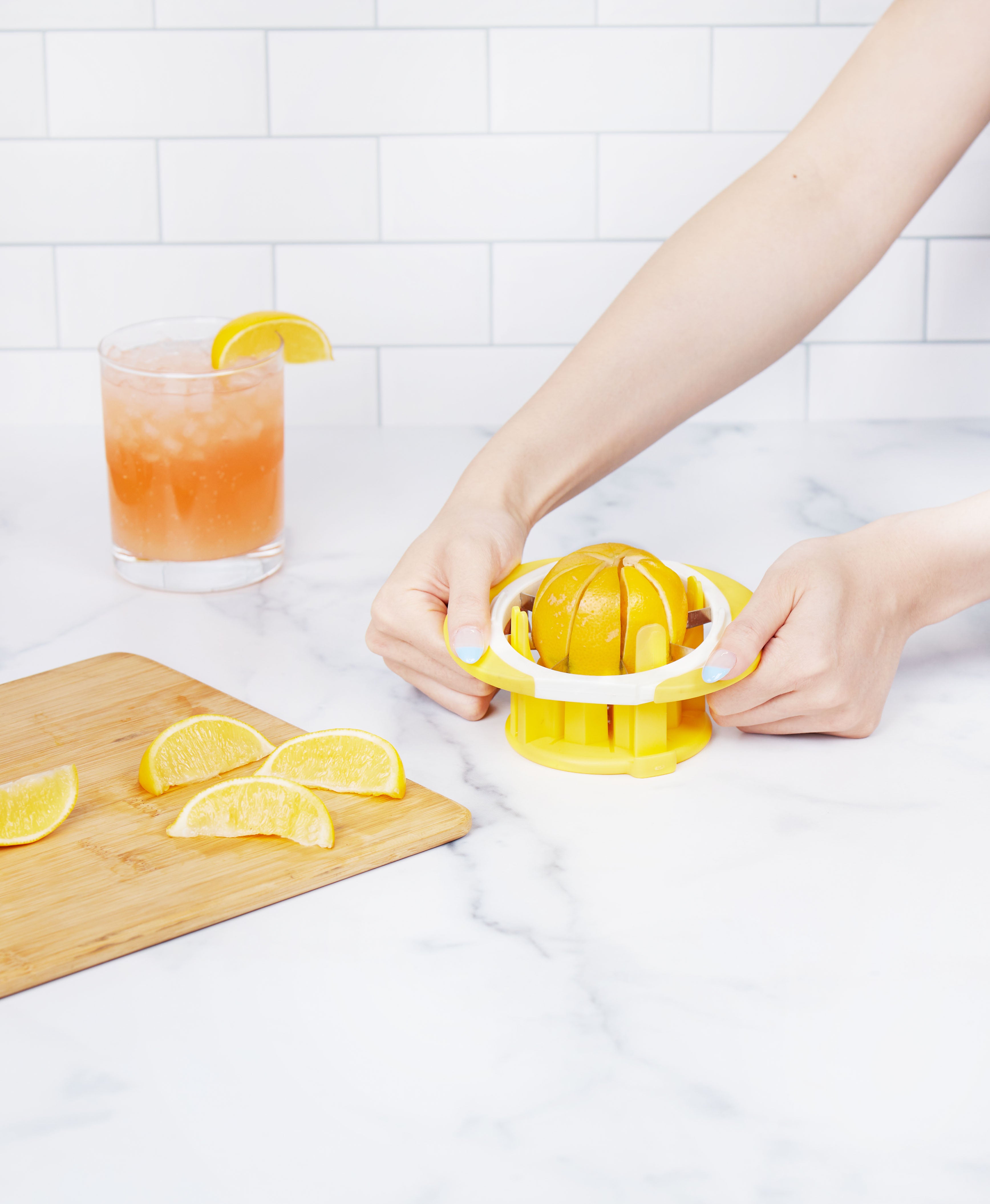 Using the yellow Citrus Slicer and Wedger, a person presses a lemon half on a marble counter; nearby, stainless steel blades cut wedges on a wooden board, with a glass of pink drink and lemon slice in the background.