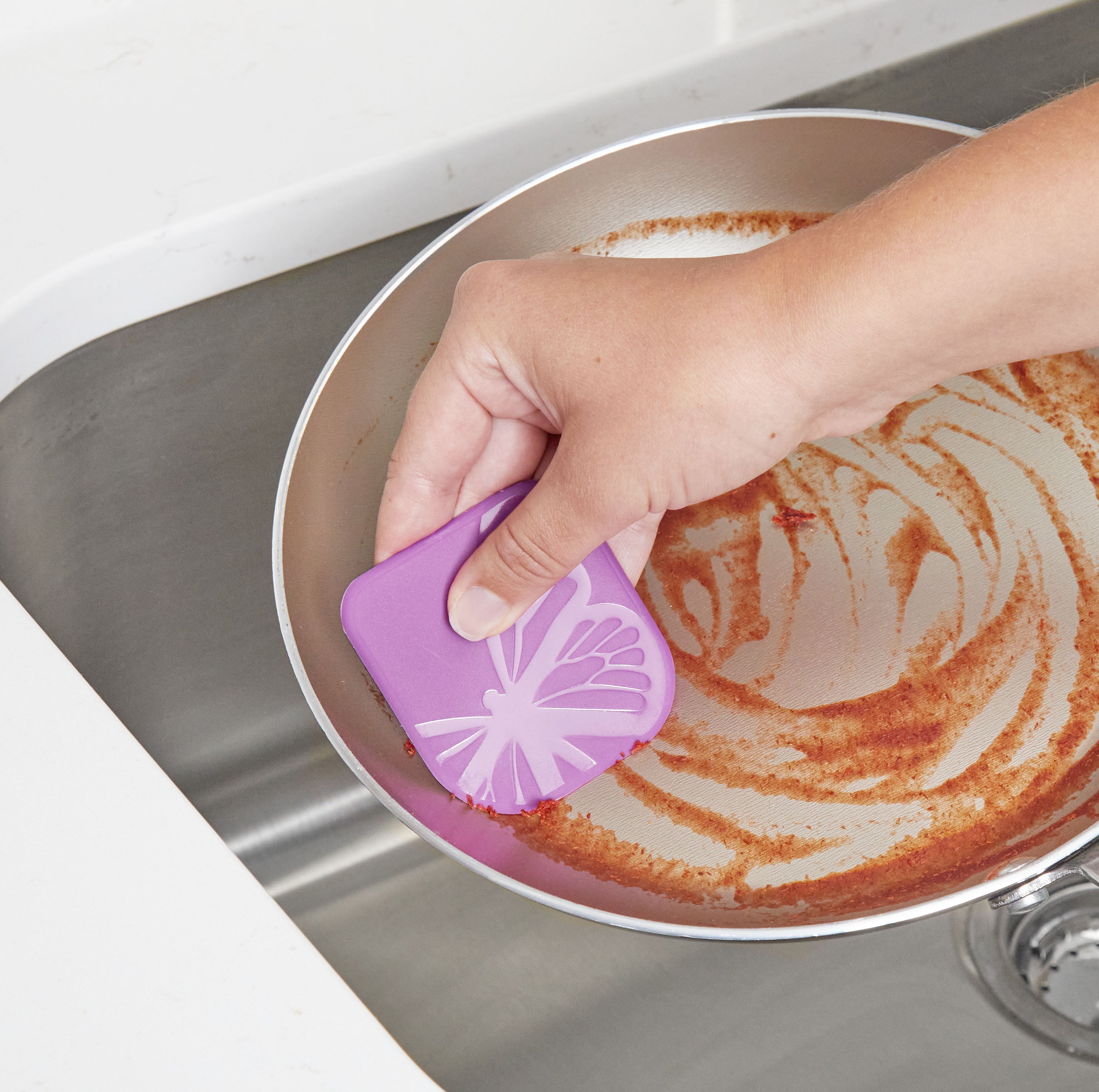 A hand uses Pan Scrapers to clean brown residue from a frying pan over a stainless steel sink, removing stains from cookware like cast iron skillets and non-stick bakeware with circular scrubbing motions.