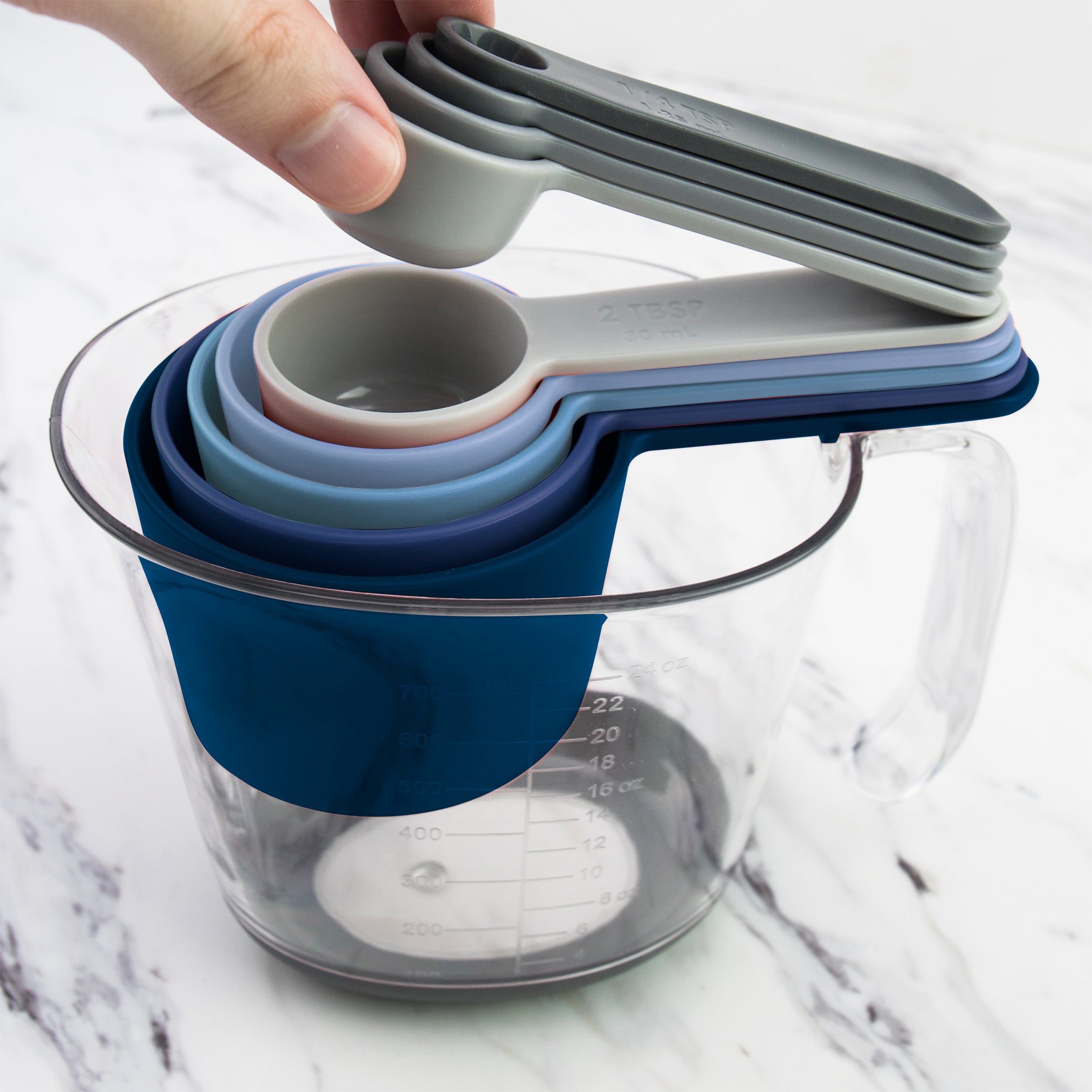 A hand lifts a gray etched spoon from the Magnetic Nested Measuring System, featuring colorful cups and spoons stacked inside a clear glass measuring cup on a white marble surface.