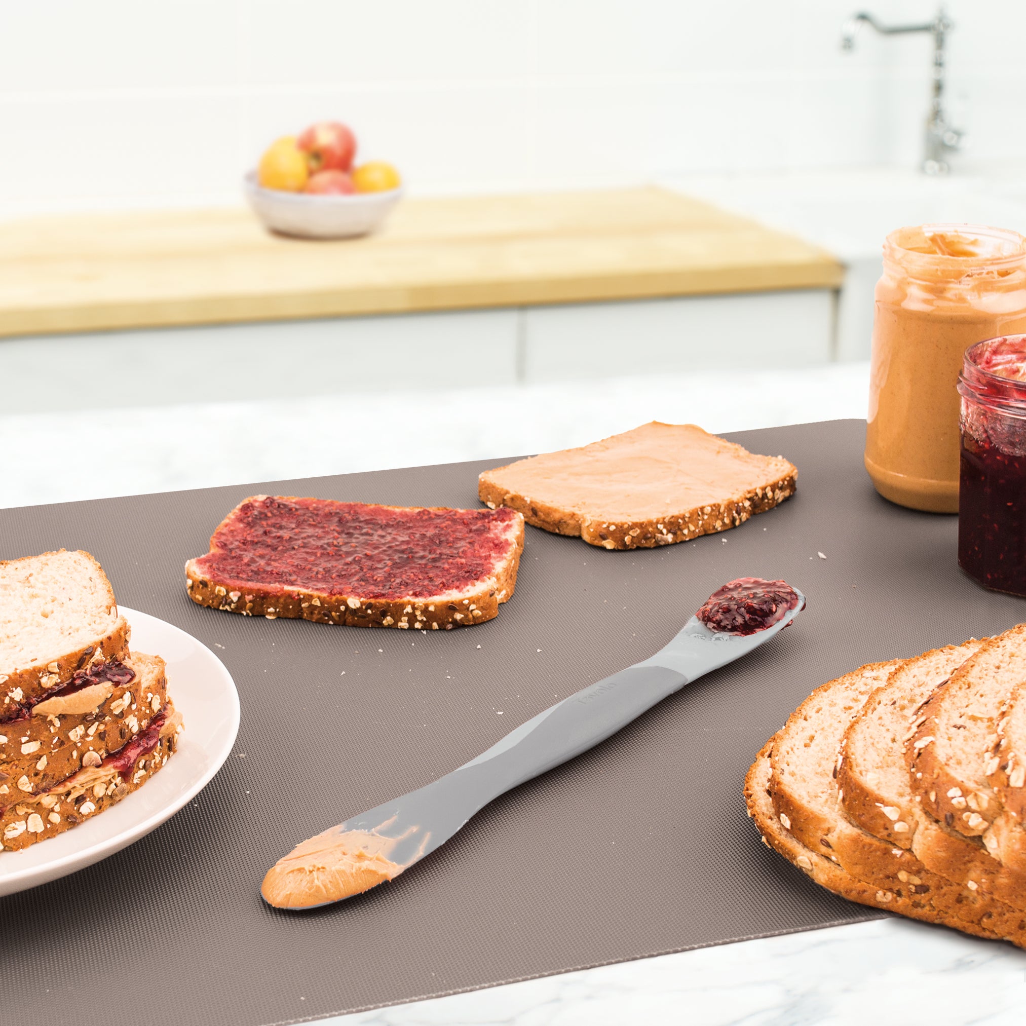 On the kitchen counter are jars of peanut butter and jelly, whole grain bread with spreads, a sandwich on a plate, and the Scoop and Spread kitchen tool. A bowl of fruit sits in the background.